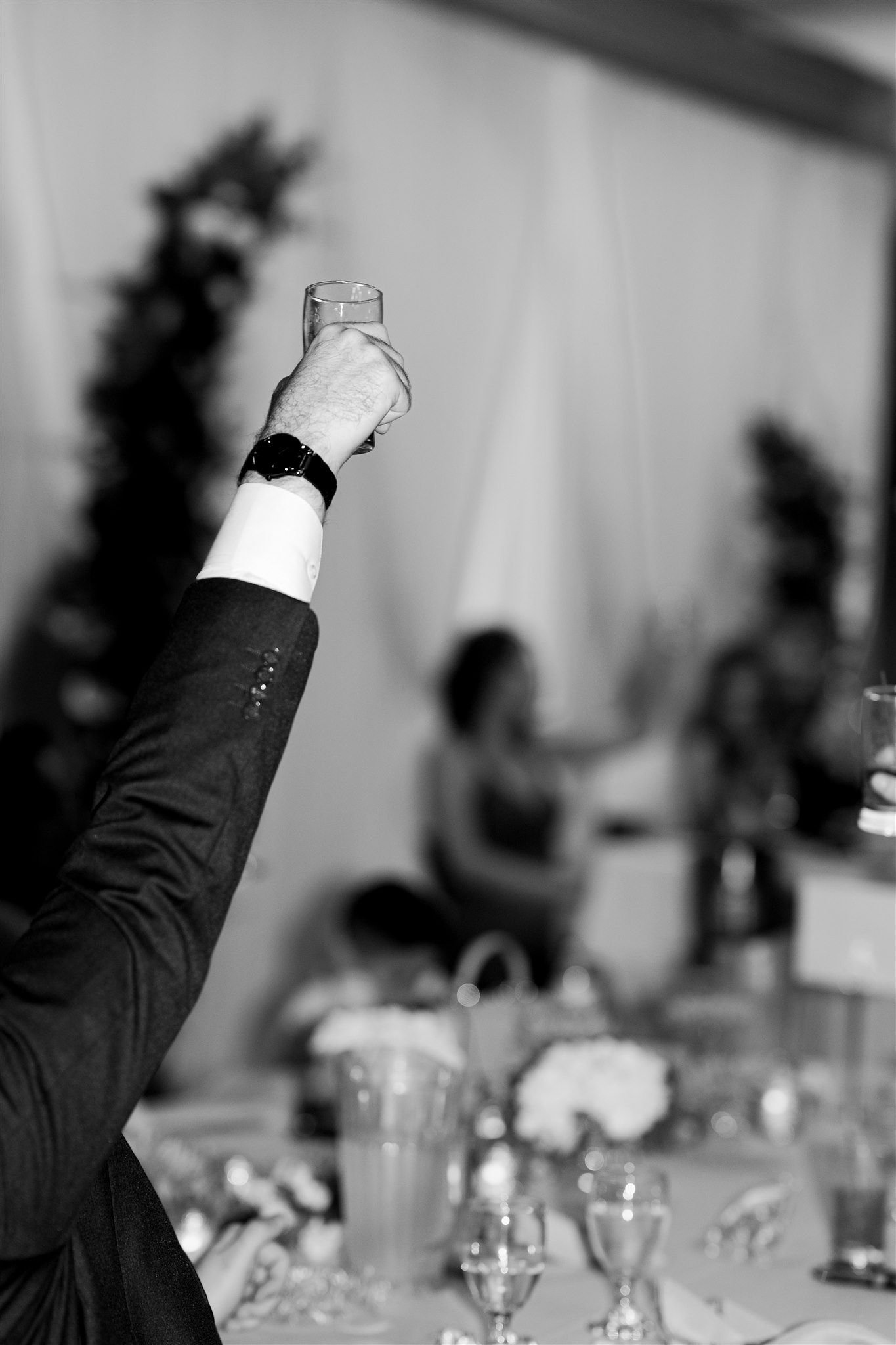 A person in a suit holding a glass of champagne raised in a toast at a celebration or party. The background shows blurred people and decorated tables in a festive setting.