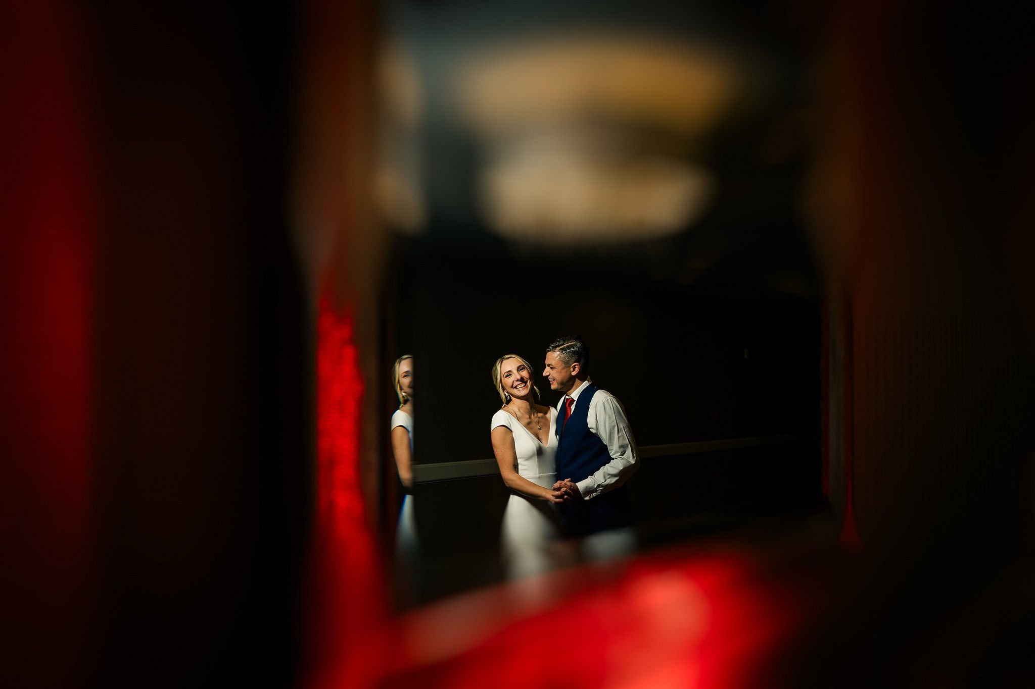 A smiling bride and groom holding hands, seen through a blurry red-framed mirror, during their wedding photos.