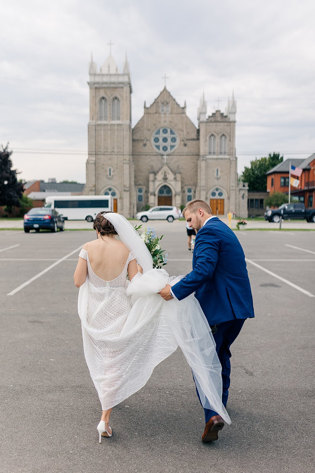 A bride and groom walking in a parking lot in front of a church, with the groom holding the bride's dress.