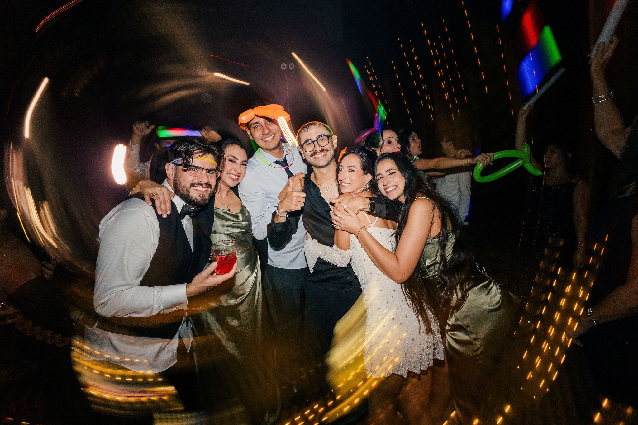 Group of people celebrating at a party, smiling and hugging, with colorful lights and glow sticks in the background.