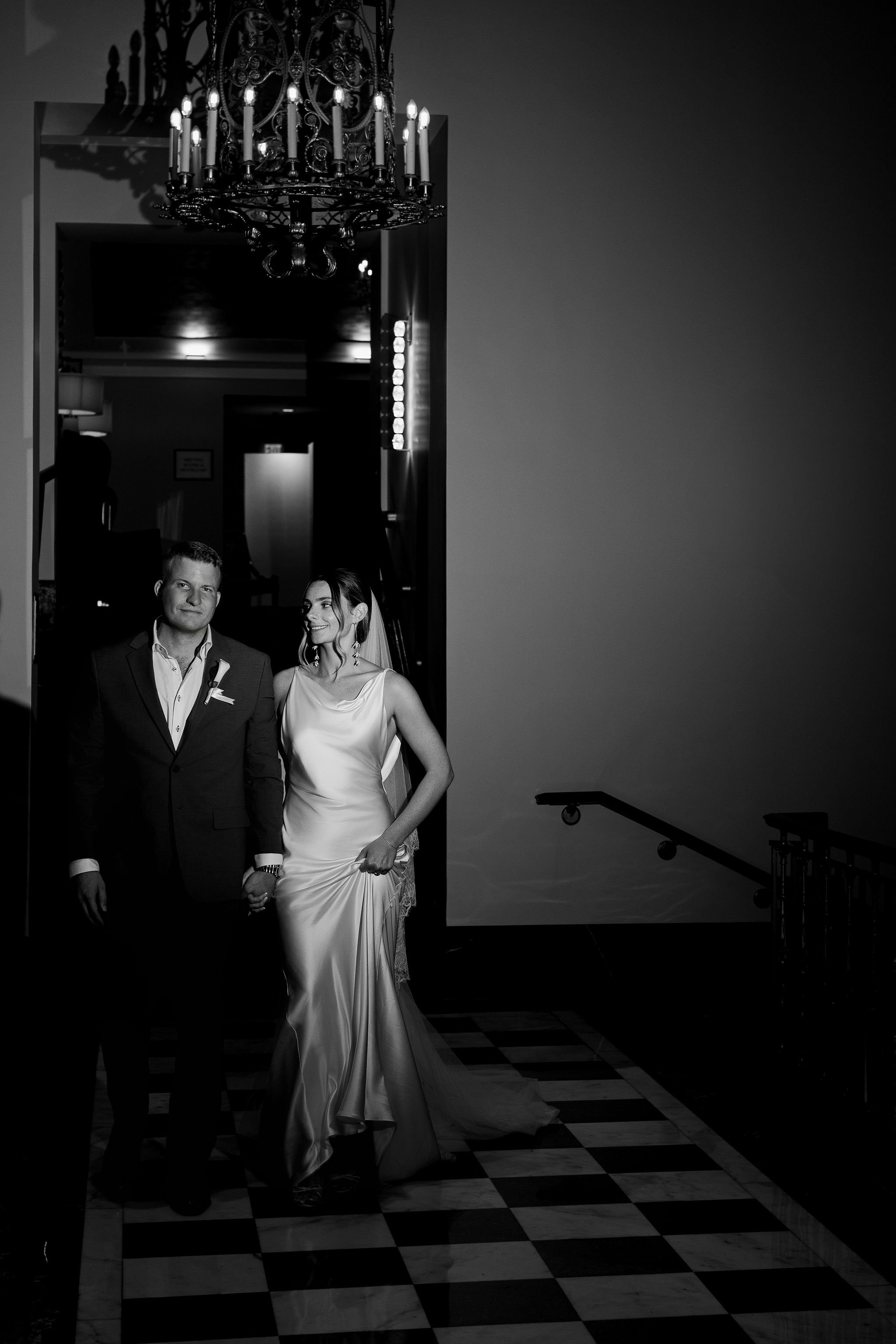 A bride and groom walking hand in hand in a dimly lit venue, with a chandelier overhead and checkered floor beneath them.