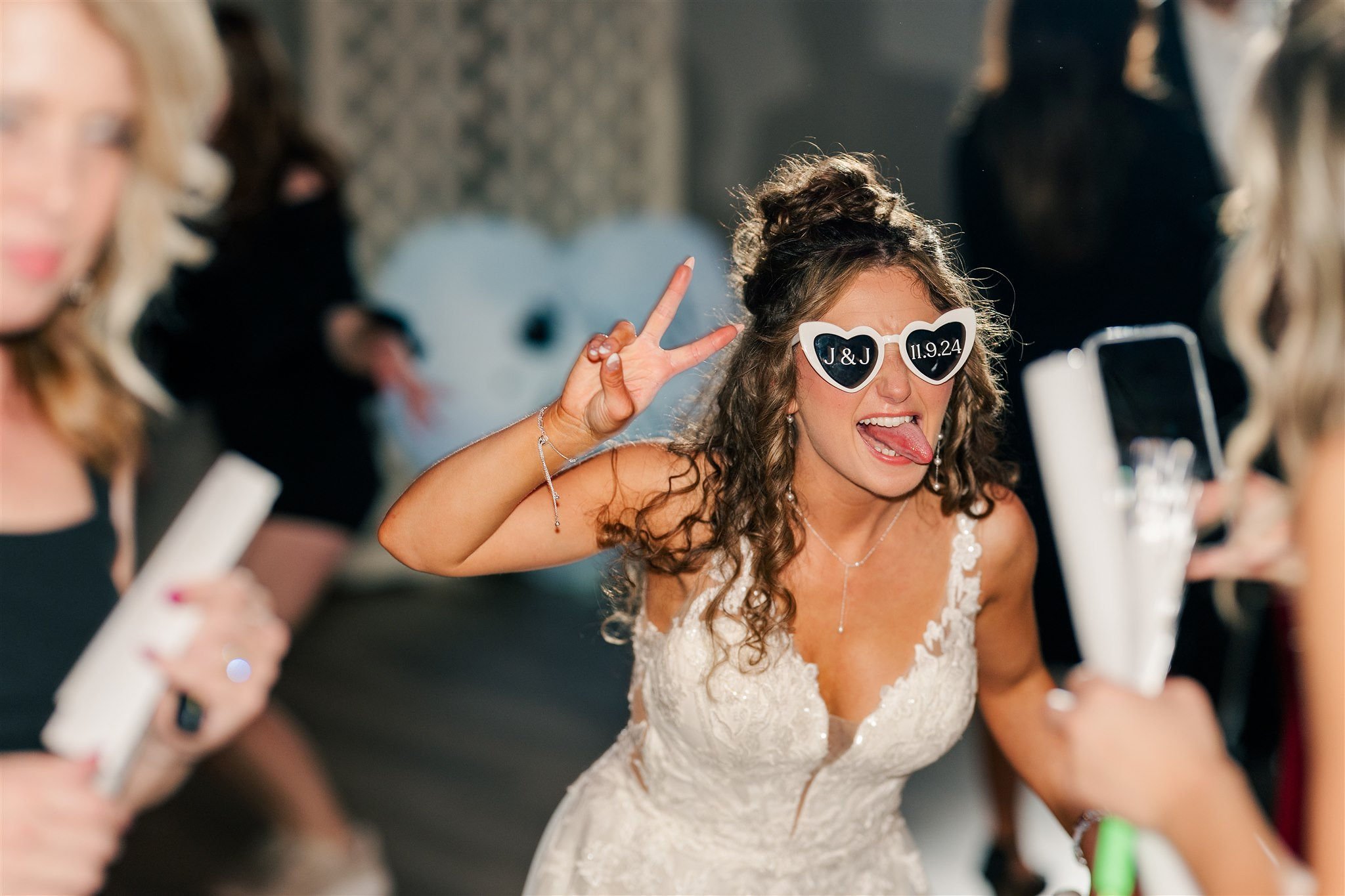 A woman in a wedding dress wearing heart-shaped sunglasses, sticking out her tongue, and making a peace sign at a celebration.
