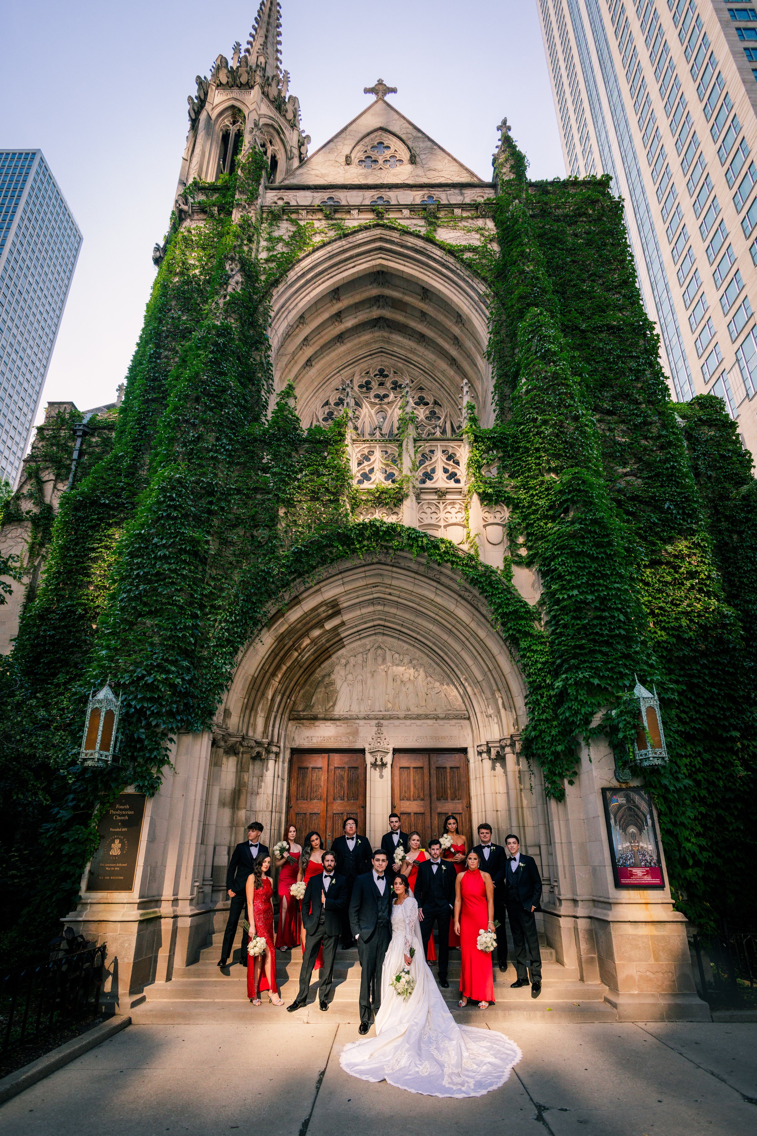 A wedding party, including a bride in a white gown and bridesmaids in red dresses, a groom, and groomsmen in black suits, standing outside a Gothic-style church with ivy-covered walls and tall towers in an urban setting.