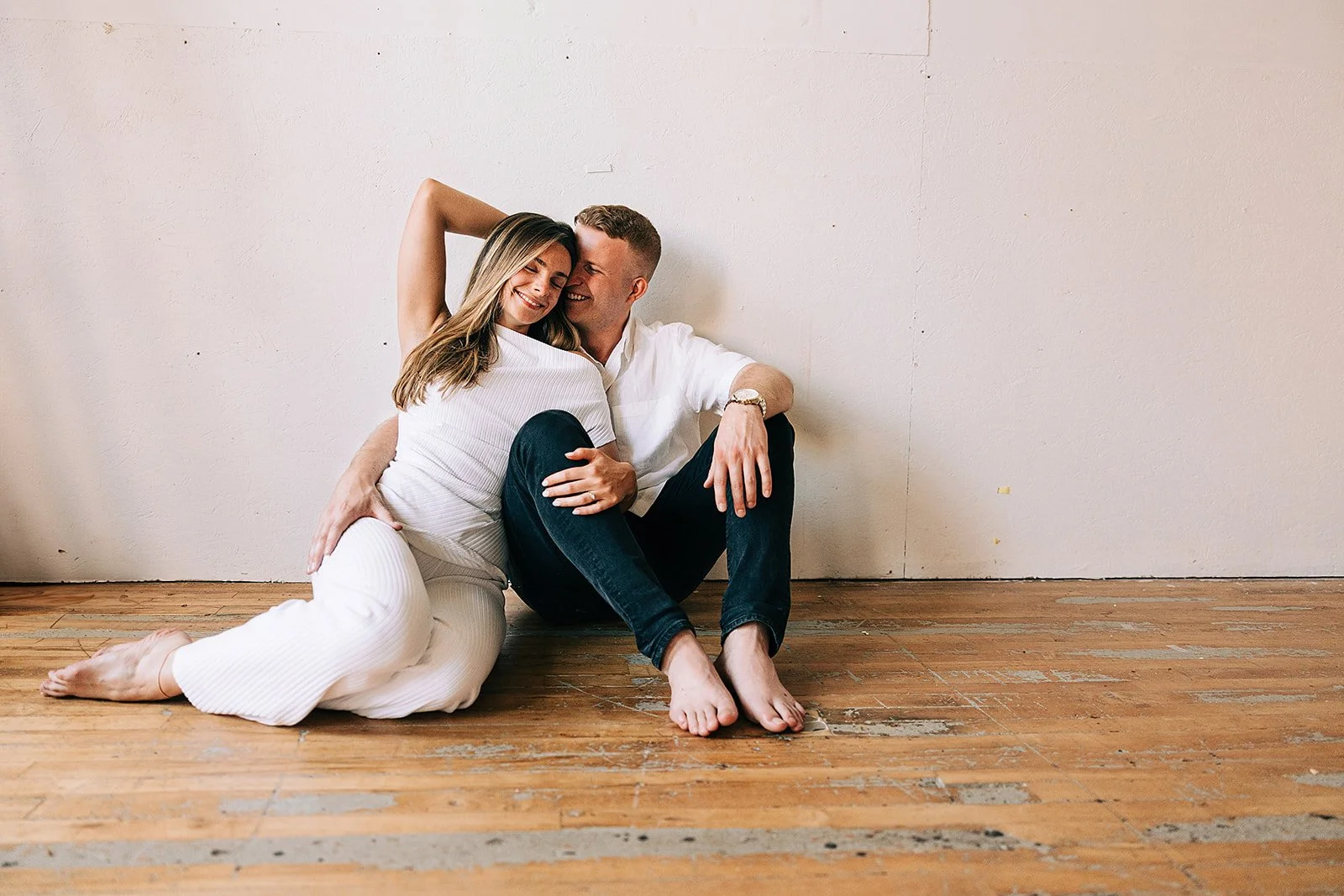 A couple sitting on a wooden floor against a plain wall, smiling and embracing each other, with the woman sitting on the floor and the man sitting with his back against the wall.