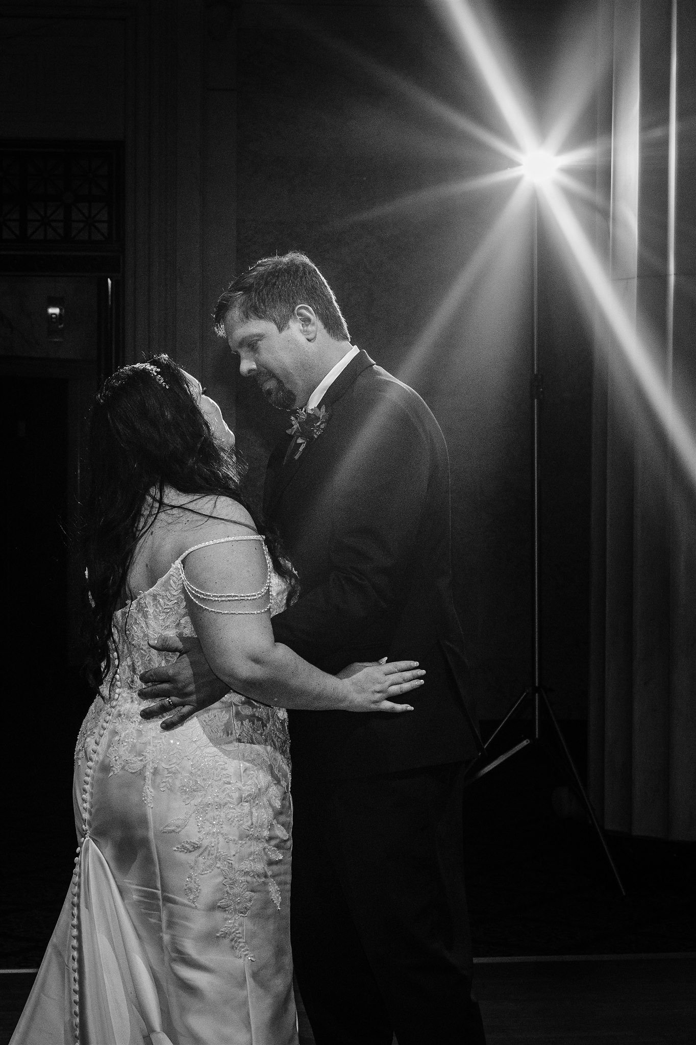 A black and white photo of a couple dancing closely, with the woman wearing a lace wedding dress and the man in a tuxedo, lit by a bright light in the background.