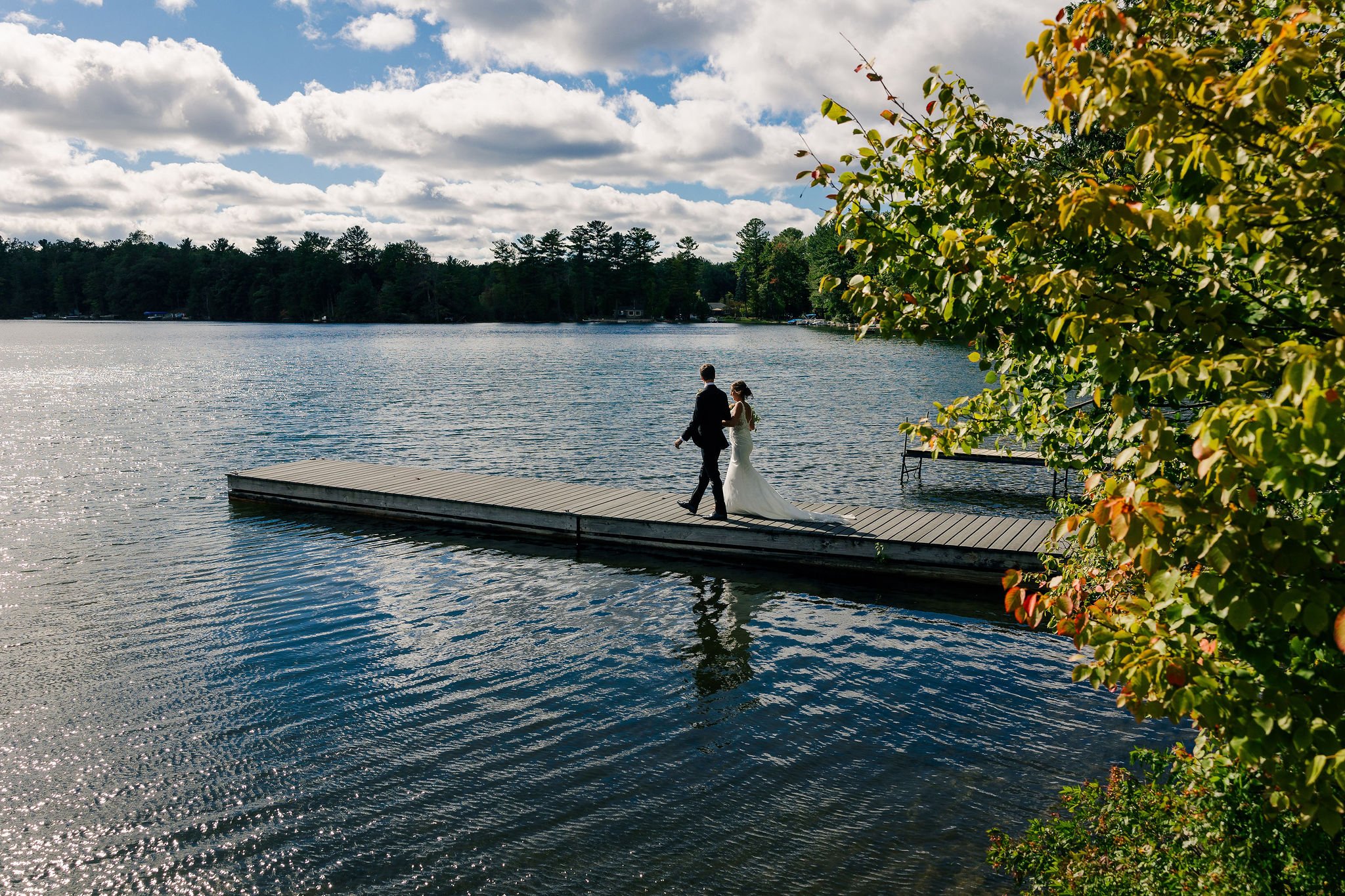 A bride and groom walk hand in hand on a dock over a lake, with trees and a cloudy sky in the background.