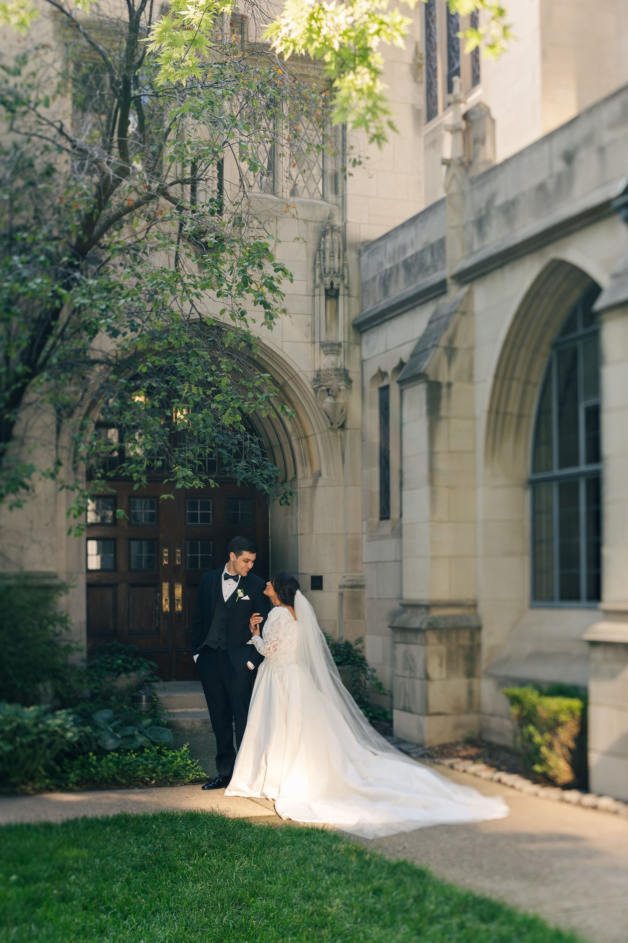A bride and groom standing outside a historic stone building with gothic architecture, smiling at each other, surrounded by greenery and trees.