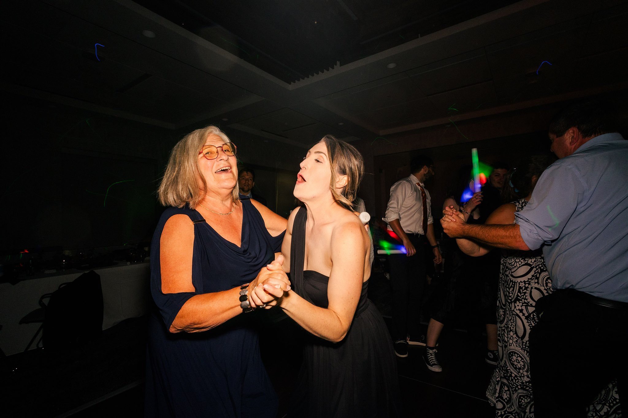 Two women dancing happily together at a party, surrounded by other dancing guests with colorful lights.