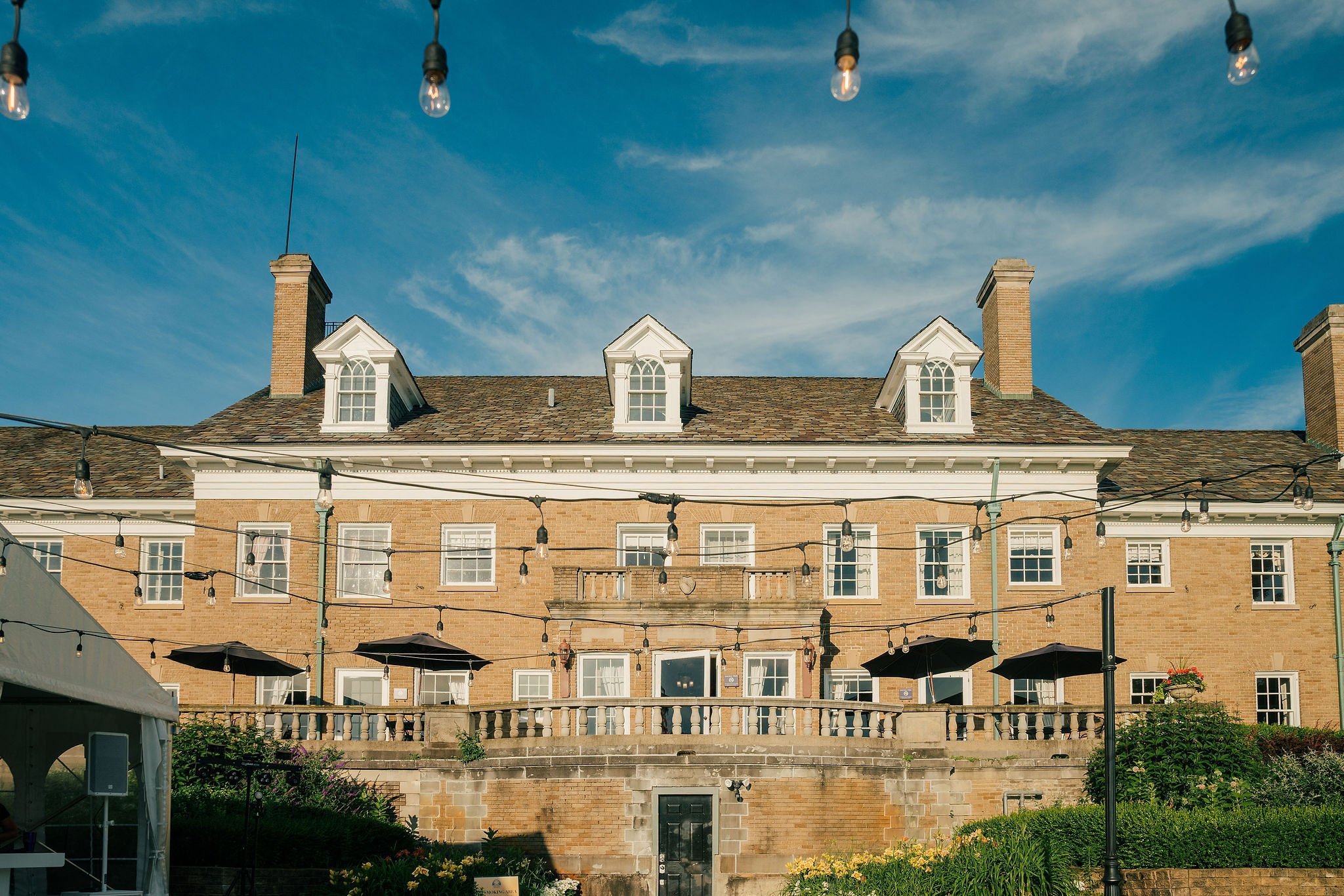 A large, three-story brick building with a sloped roof, multiple chimneys, and dormer windows under a blue sky with wispy clouds. String lights hang across an outdoor patio area with umbrellas and lamp posts.