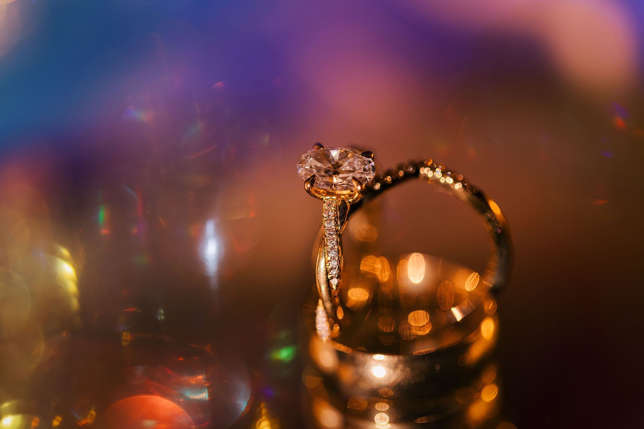 Close-up of a diamond engagement ring with a large center stone and a wedding band with smaller diamonds, all set in gold, with colorful bokeh background.