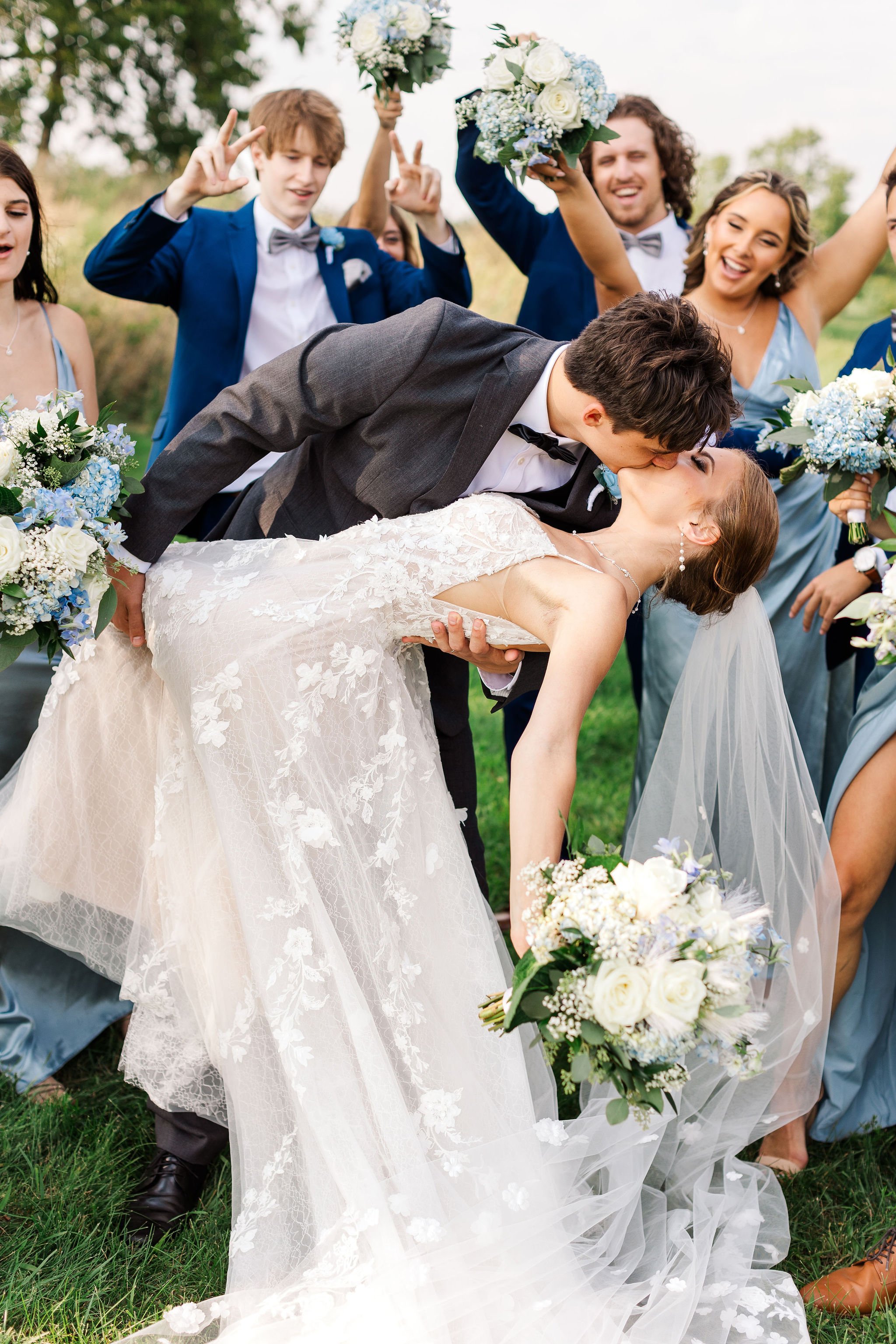 Bride and groom kissing during wedding photo with bridal party around them, holding bouquets, outdoors.