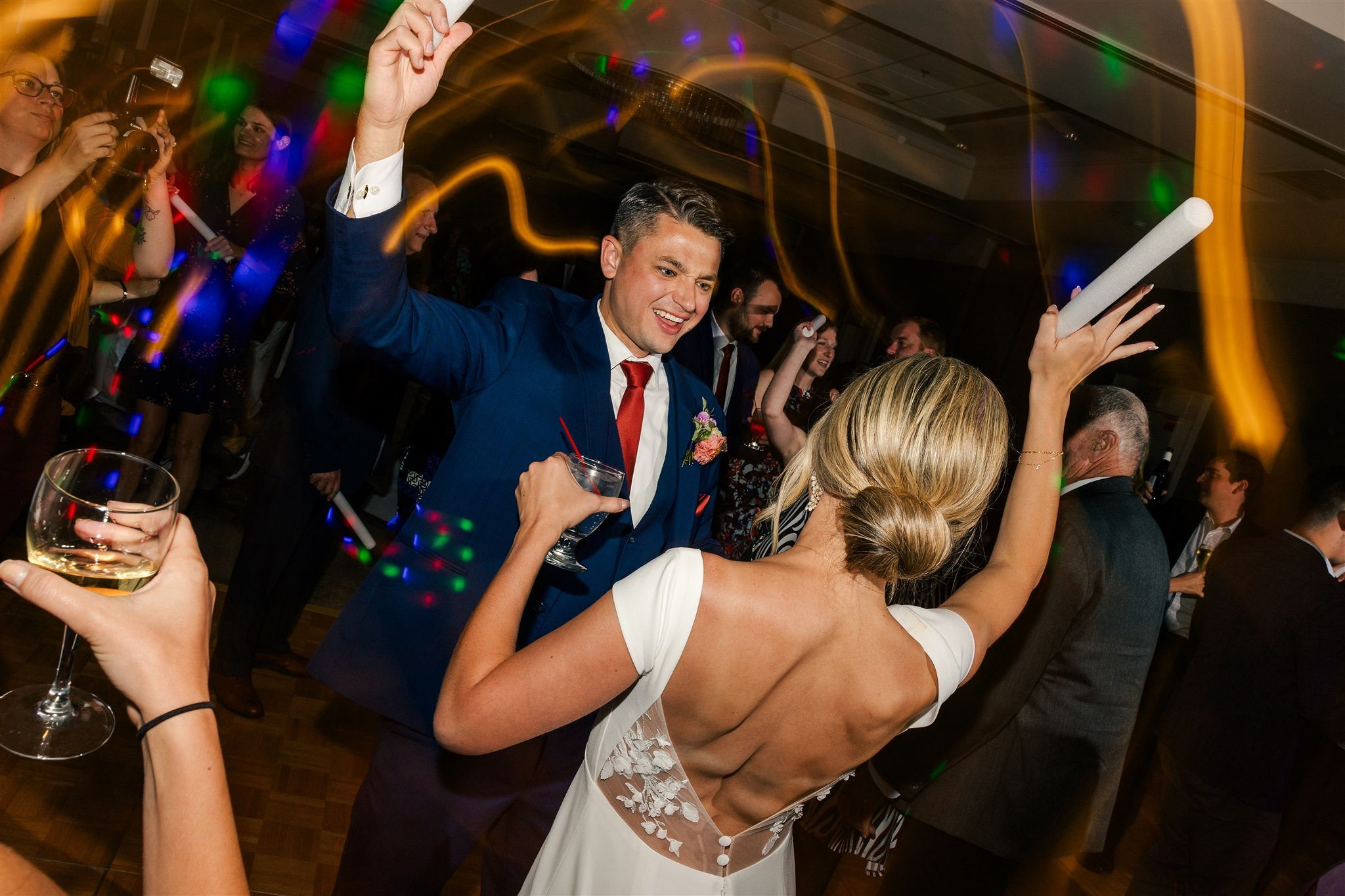 People dancing and celebrating at a wedding reception with colorful lights and drinks.