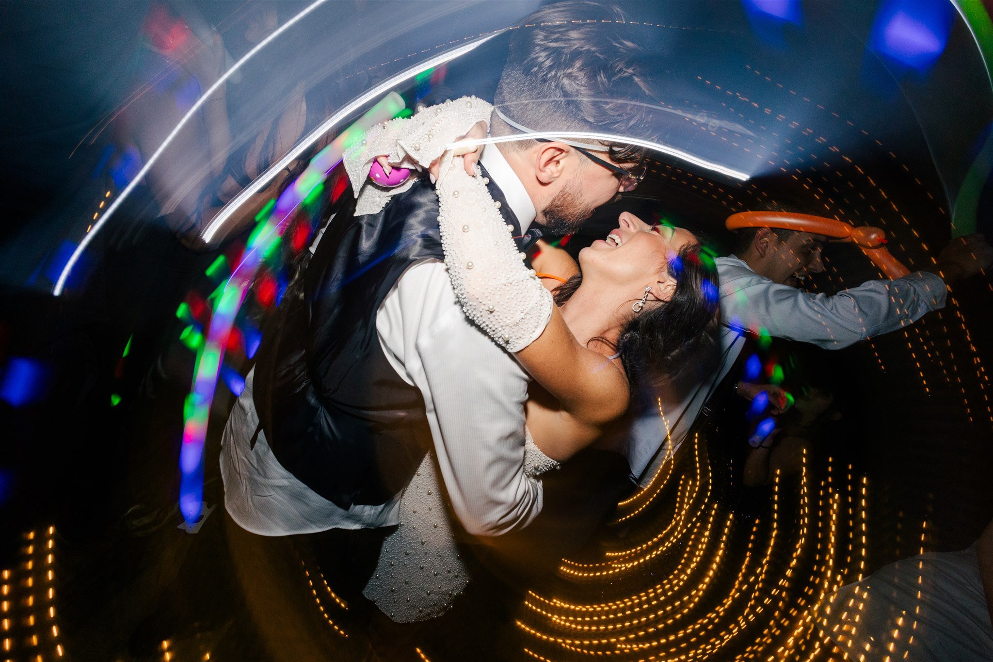 Couple dancing closely at a party, smiling and looking at each other, with colorful lights and motion effects creating a lively atmosphere.