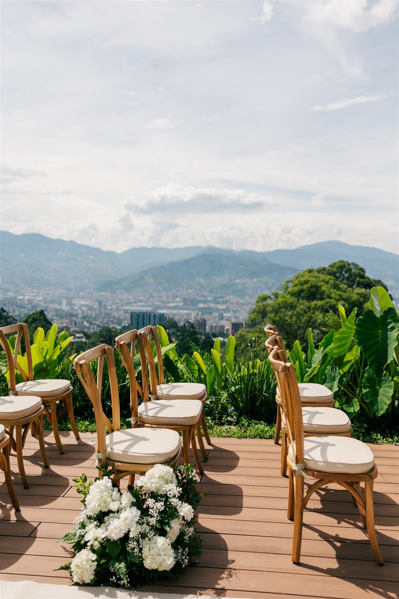 Outdoor wedding ceremony setup with wooden chairs and white floral arrangements on wooden deck, overlooking a green valley and mountains under a partly cloudy sky.