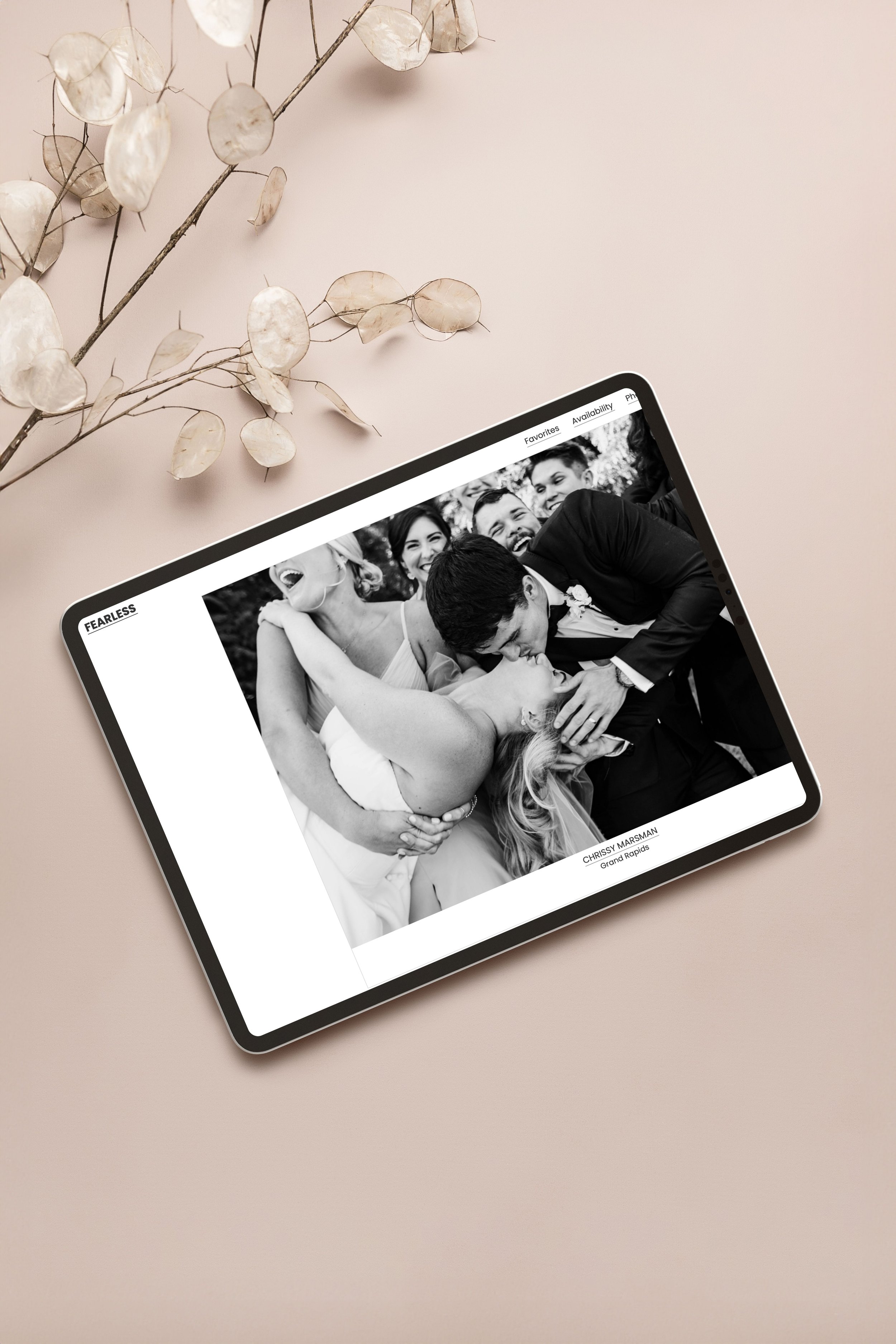 Tablet displaying a black and white wedding photo of a man and woman kissing, with several people smiling behind them, on a light pink surface next to a dried plant with round seed pods.