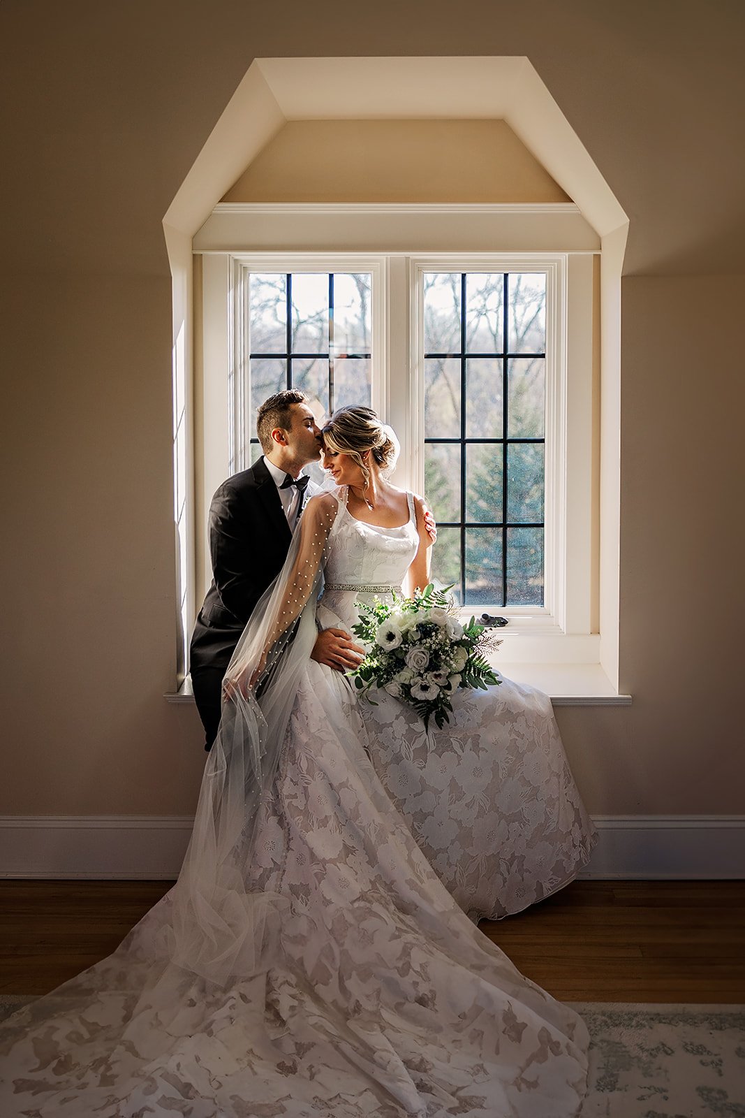 A bride and groom sit close together by a window in a warmly lit room, with the groom kissing the bride's forehead. The bride holds a bouquet of white flowers, wearing a white lace wedding dress with a long train, and a sheer veil. The groom is dress