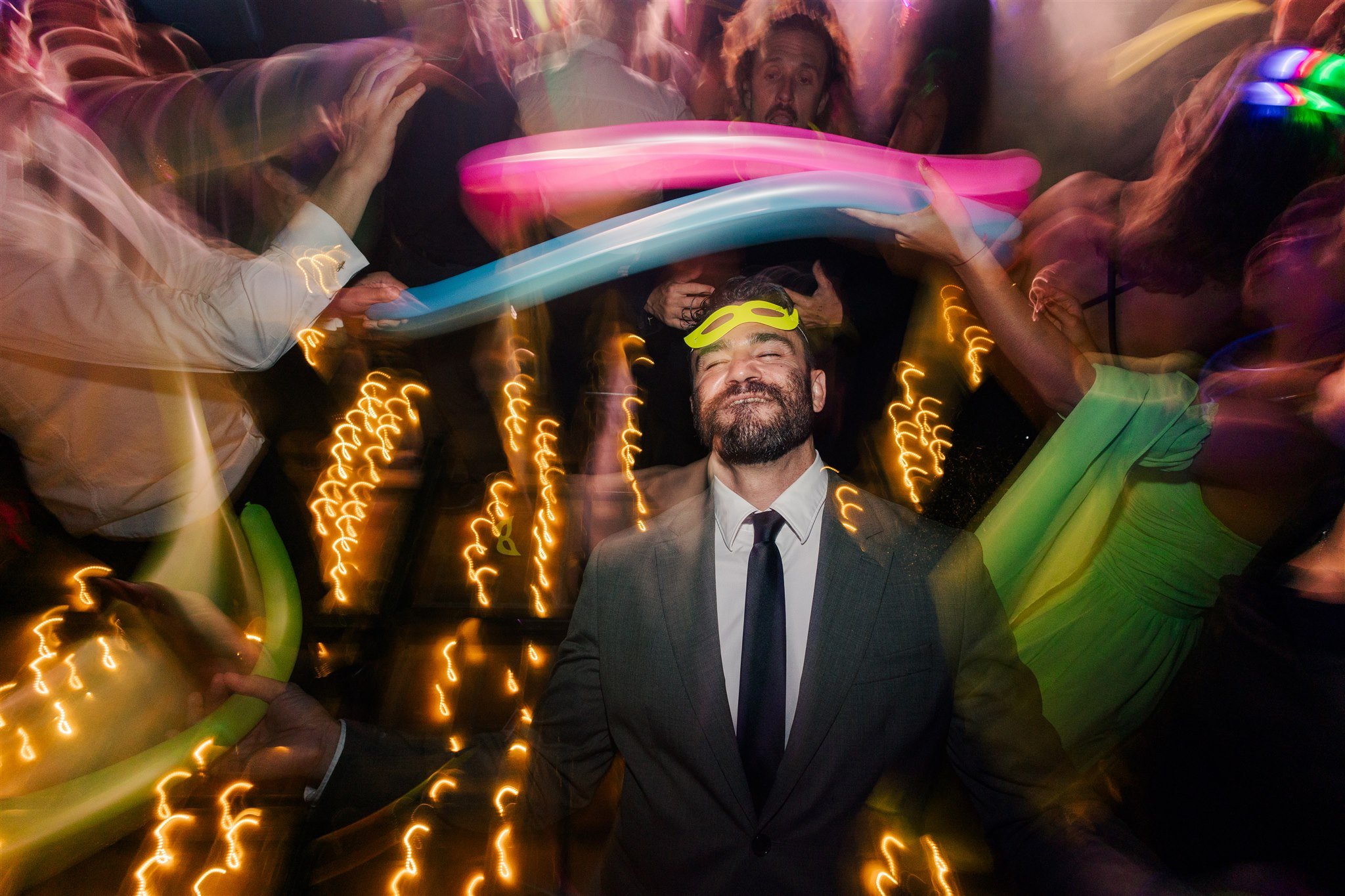 A man in a suit and tie wearing glow glasses smiling with eyes closed at a party, surrounded by people dancing and light streaks from colorful balloons and lights.