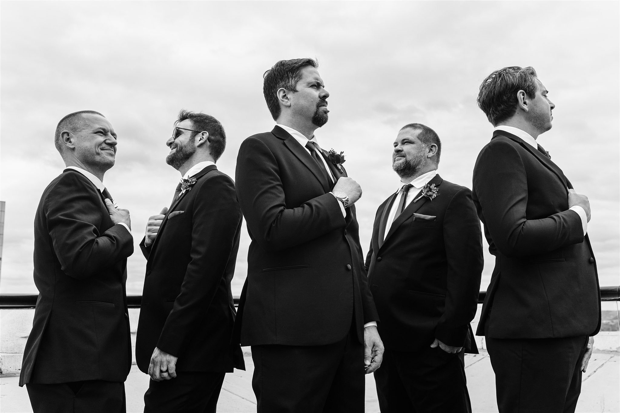 Five men in tuxedos stand outdoors against a cloudy sky, with their right hands over their hearts, looking in different directions.