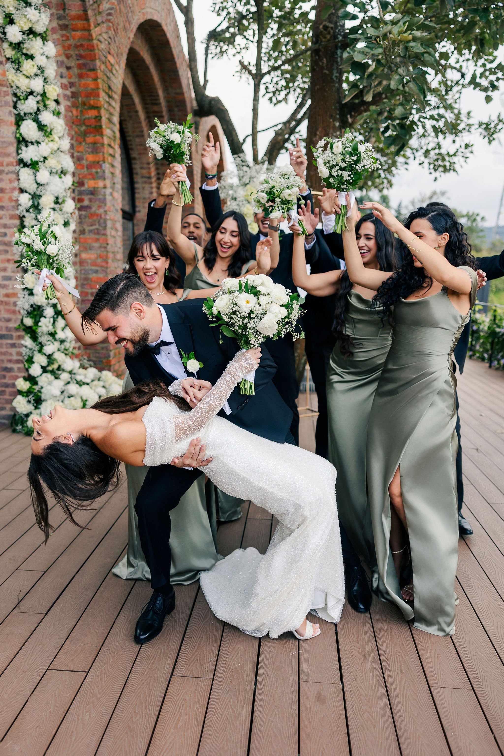 A joyful wedding celebration with a bride and groom, surrounded by bridesmaids and groomsmen holding bouquets, as the groom dips the bride for a dance or photo.