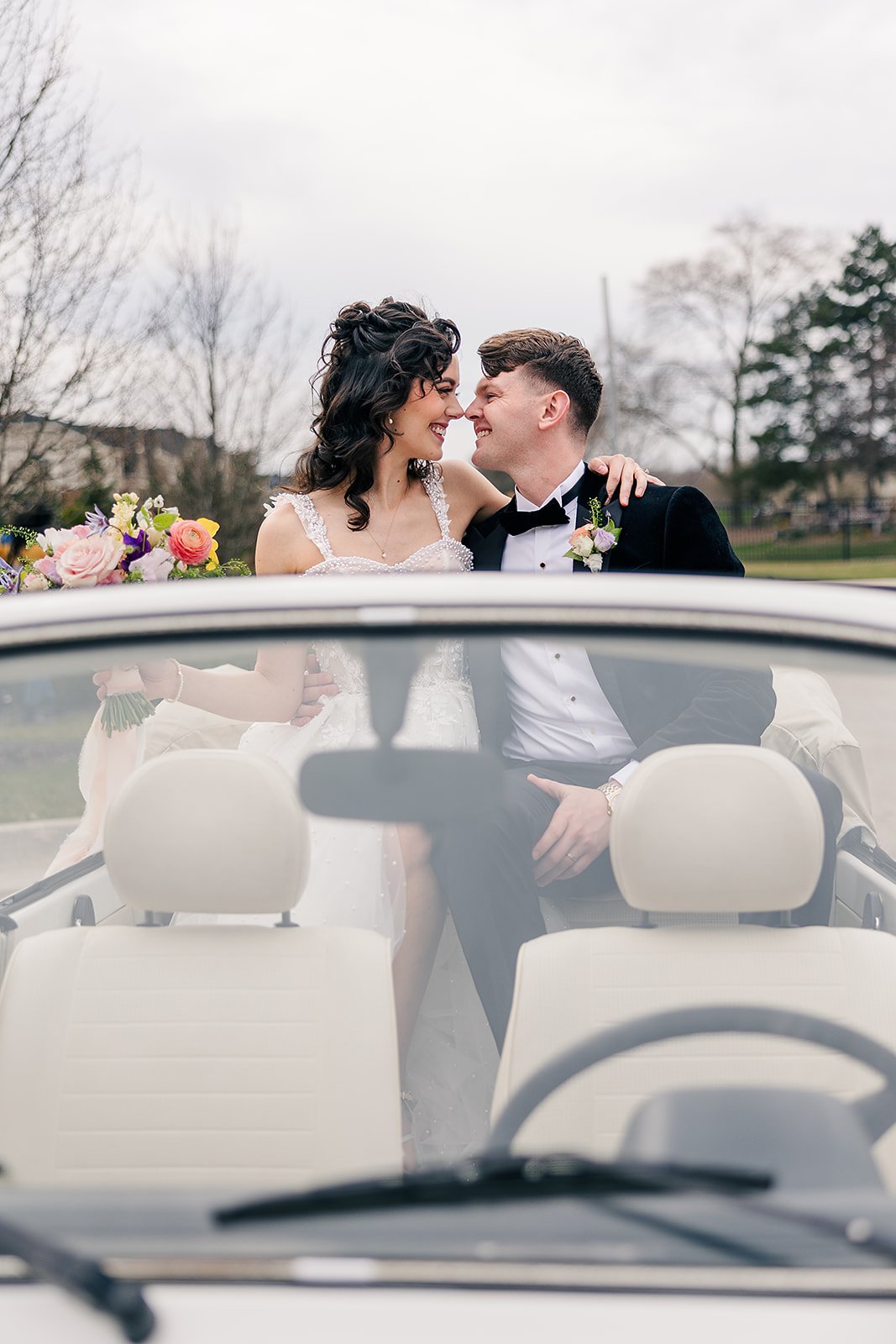 A newlywed couple in wedding attire sitting in a car, smiling and leaning their foreheads together, with a bouquet of flowers visible on the left side.