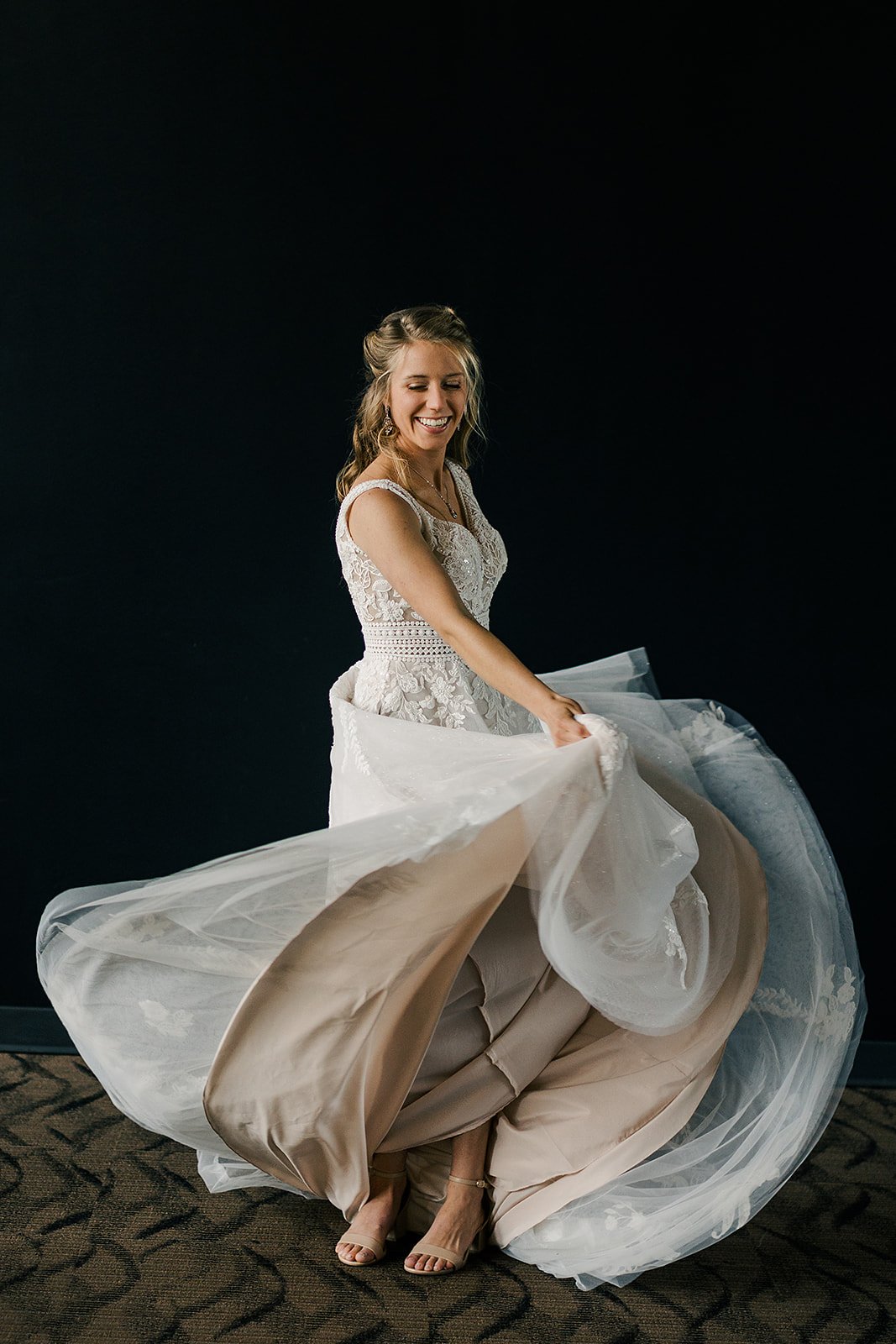 A woman in a bridal gown smiling and twirling, lifting the layers of her wedding dress to reveal her shoes against a black background.