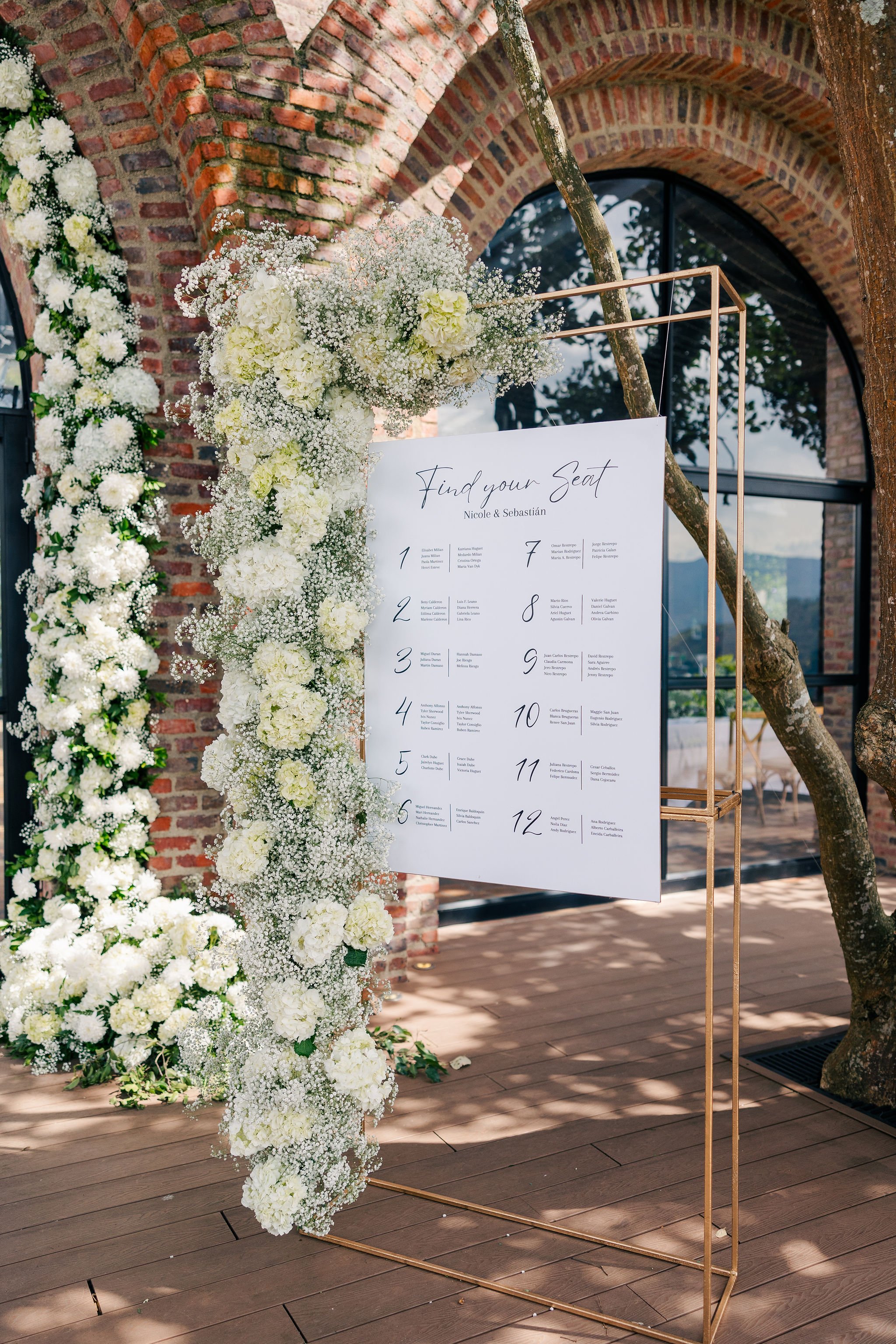 A wedding seating chart display framed with a gold metal stand, decorated with white floral arrangements including baby's breath and hydrangeas, set against a rustic brick wall with a large window in the background.