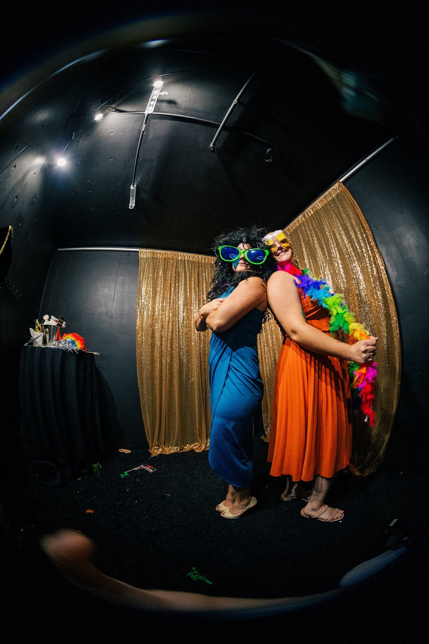 Two women in colorful outfits and fun oversized glasses pose back-to-back at a photo booth with gold curtain backdrop and party accessories.