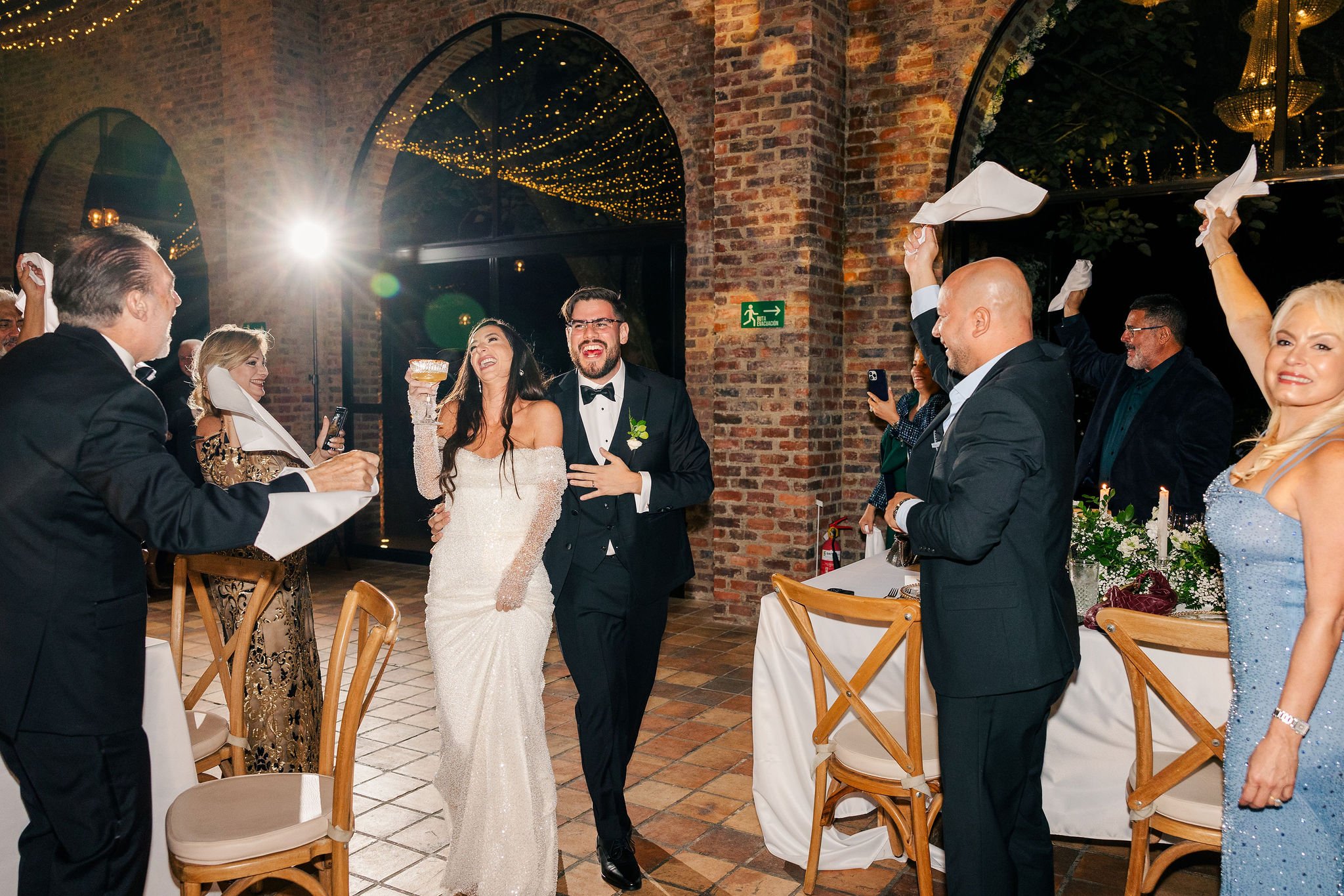 A newlywed couple, the bride in an off-shoulder white gown and the groom in a tuxedo, are celebrating at their wedding reception with friends and family. The guests are cheering and waving napkins in a festive indoor venue with brick walls and string