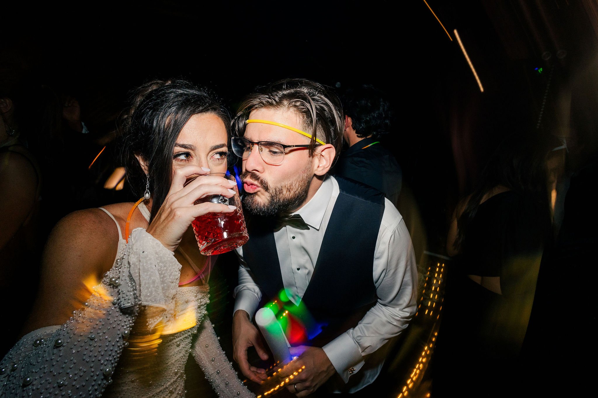 Man and woman dancing at a party, the woman is drinking from a red glass, with dark background and colorful lights.