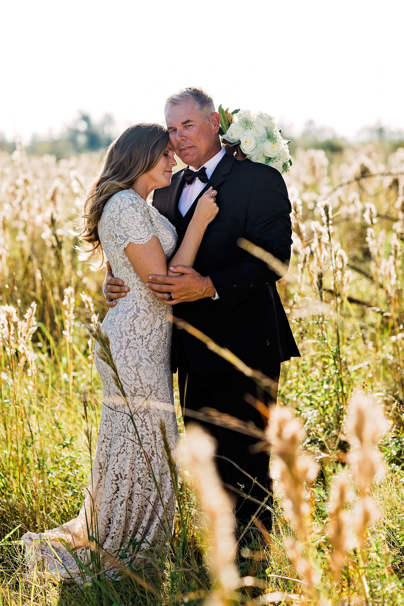 A bride and groom embrace in a field of tall grass, with the groom holding a bouquet of white flowers behind him. The bride is wearing a lace dress, and the groom is in a black tuxedo with a bow tie. They appear to be at their wedding, sharing an int