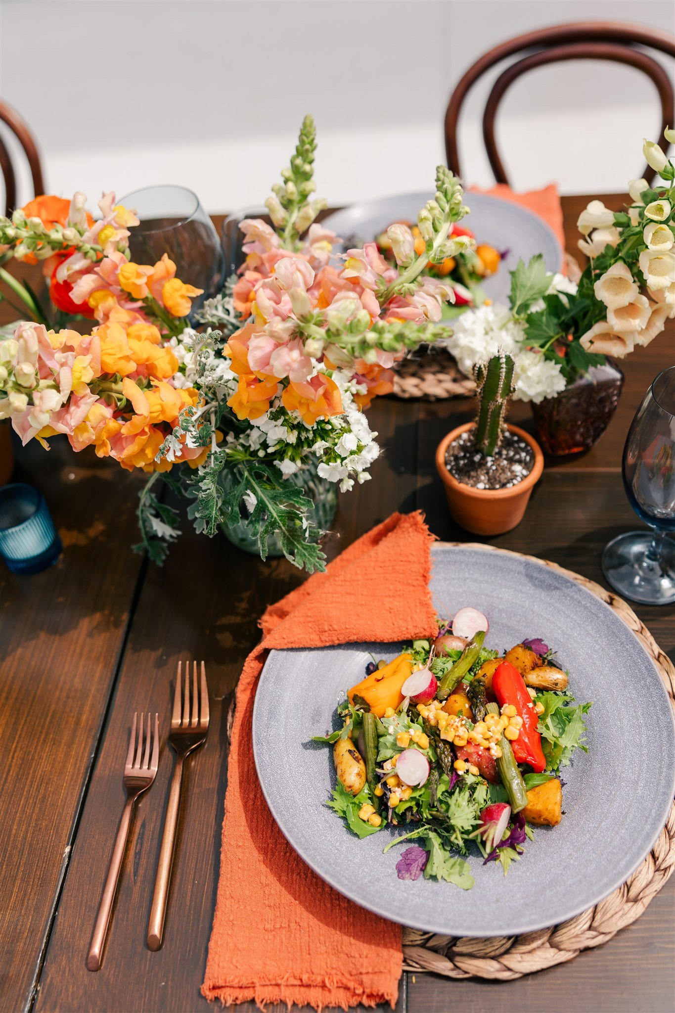 A beautifully set dining table with a colorful vegetable salad on a gray plate, orange napkin, surrounded by vibrant flowers including snapdragons, and a cactus in a terracotta pot.