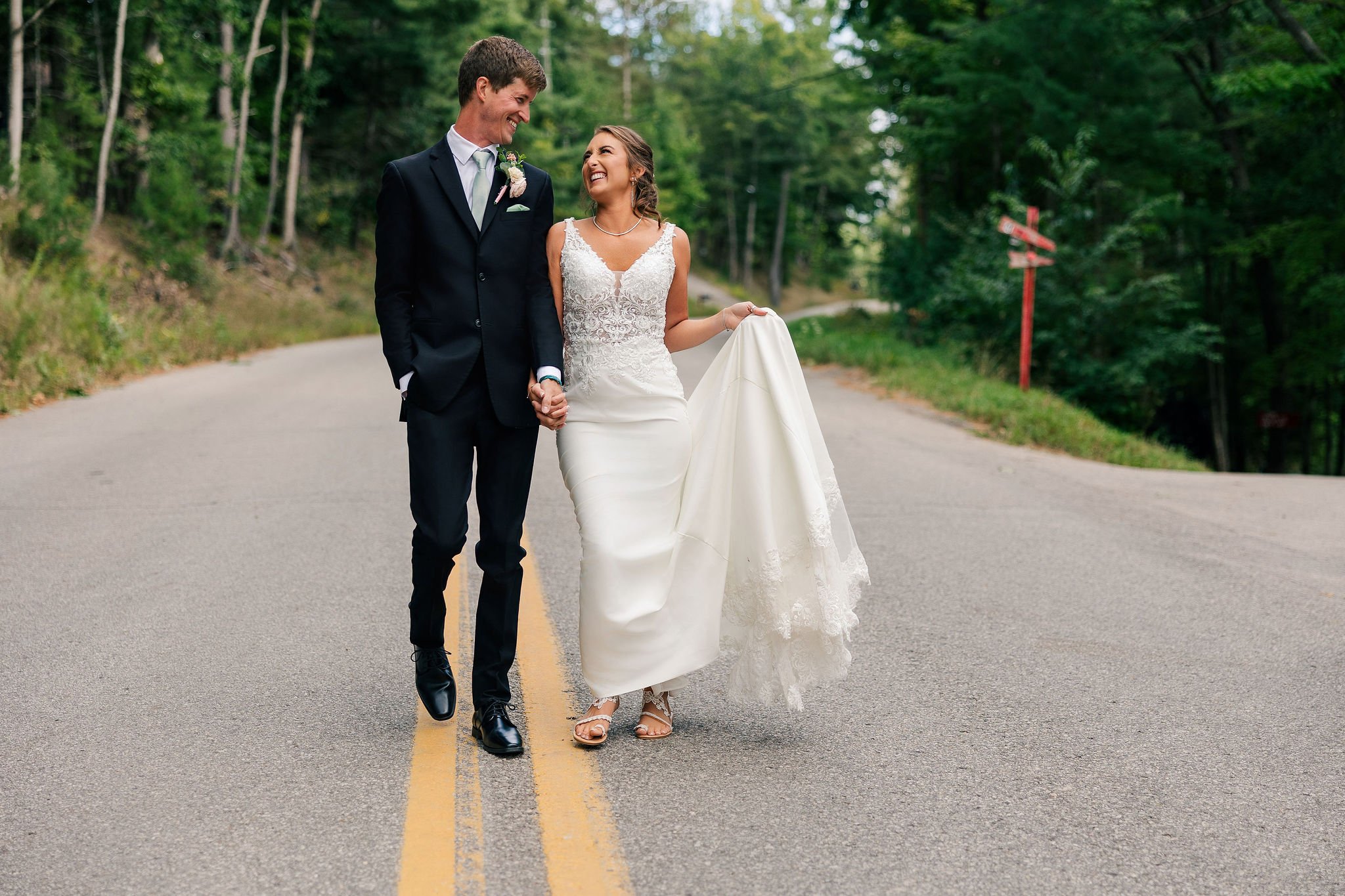 A bride and groom walking hand in hand on a rural road surrounded by greenery.