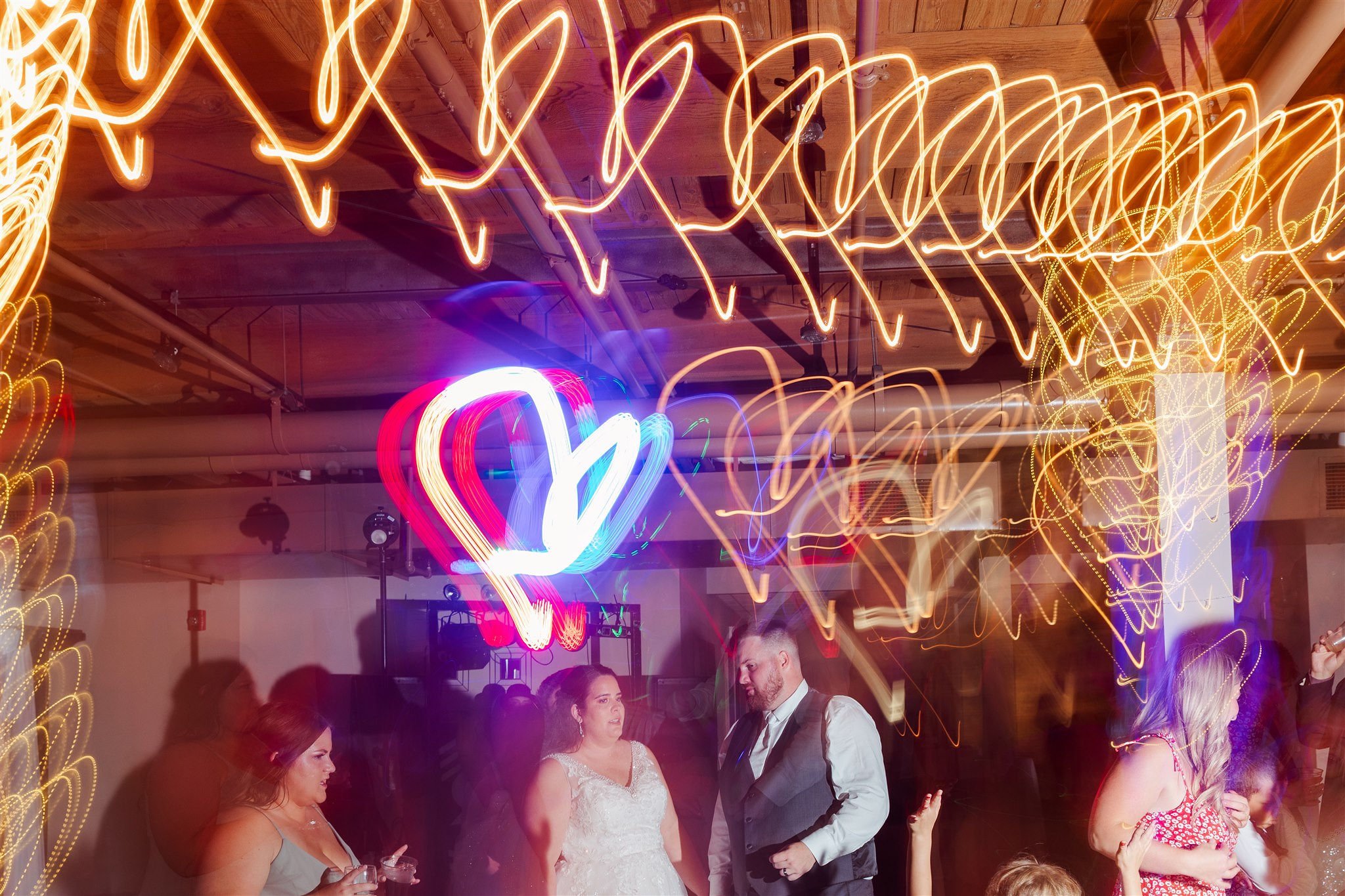 People dancing and socializing at a wedding reception with colorful neon heart decorations and string lights on the ceiling.