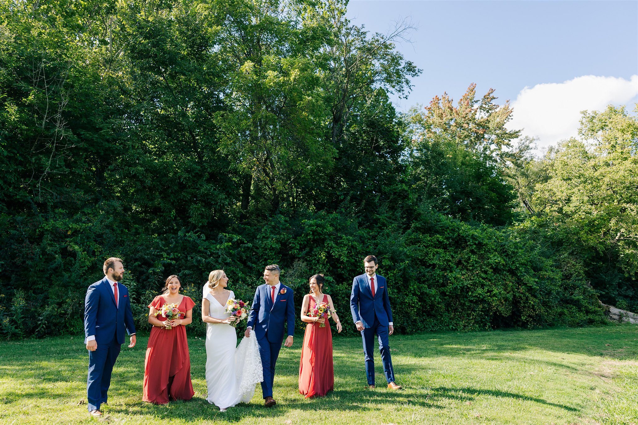 A wedding party walking outdoors on a grassy field with lush green trees in the background, including a bride in a white gown, a groom in a blue suit, and four bridesmaids and groomsmen in matching red dresses and blue suits.