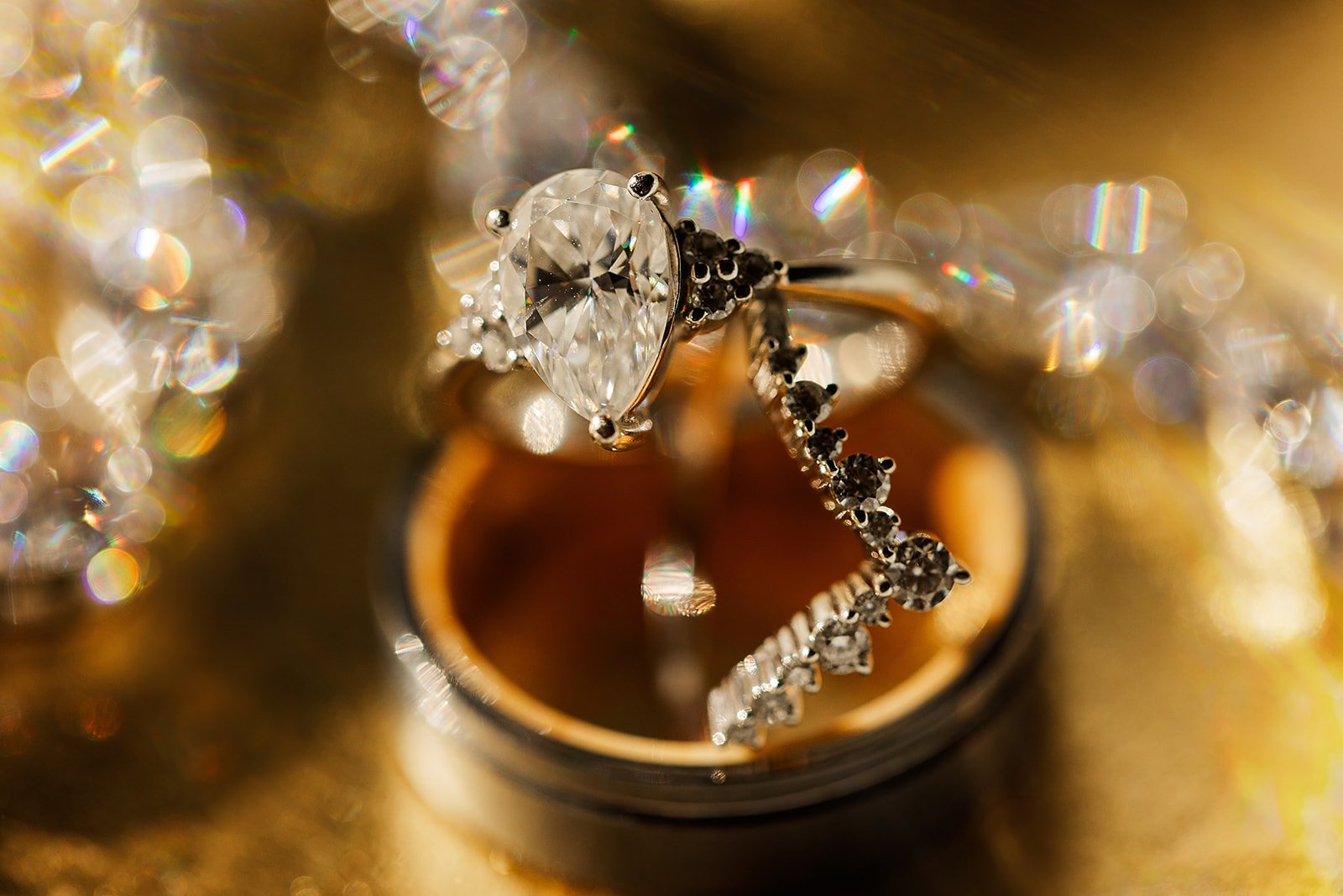 Close-up of a diamond engagement ring with a large central diamond and a wedding band with smaller diamonds, both resting on a reflective gold surface with sparkling bokeh in the background.