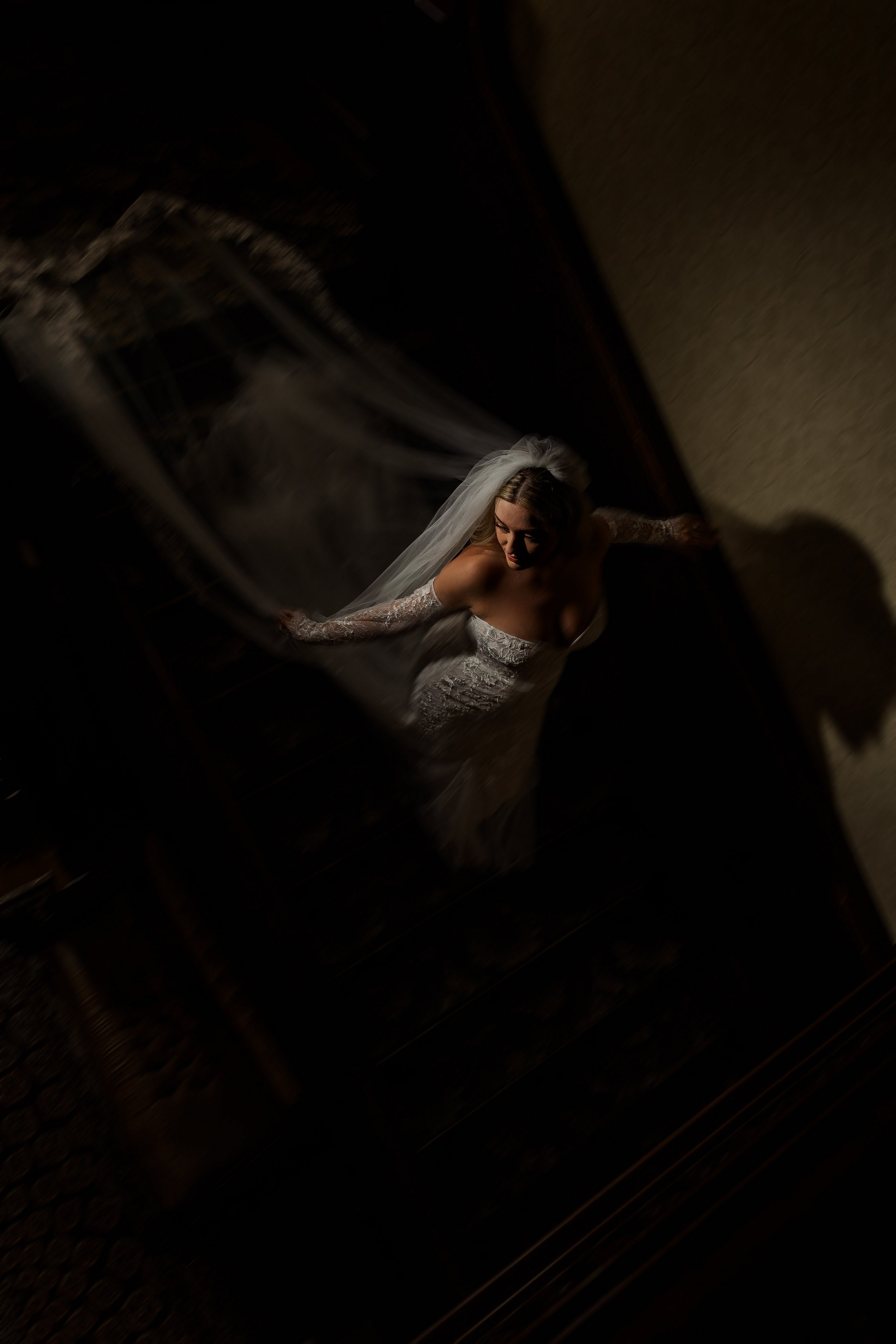 Bride wearing a white wedding dress and veil standing on a stairway, viewed from above in a dimly lit room.