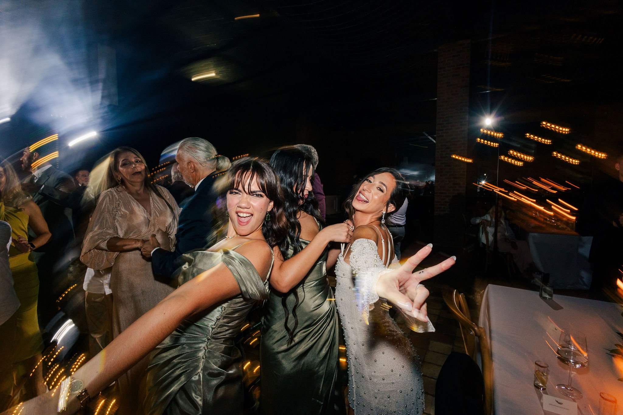 Group of women dancing and celebrating at a party or wedding reception, with blurred motion and colorful lights in the background.