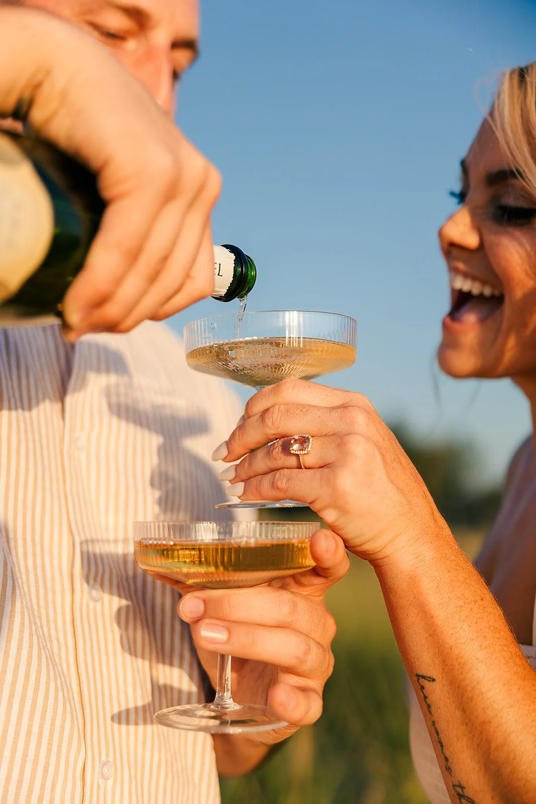 Two people celebrating outdoors with champagne glasses, one pouring champagne into the other's glass, both smiling and dressed casually, with a clear blue sky in the background.