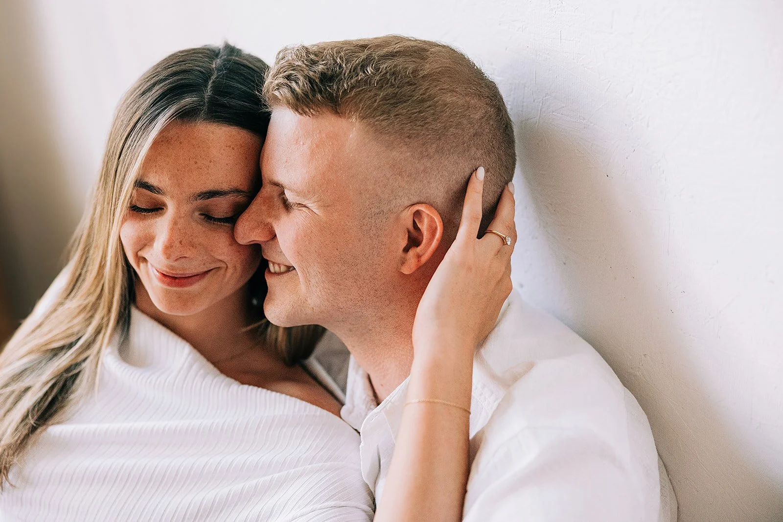 A happy couple sharing an intimate moment hugging and smiling with eyes closed, standing against a white textured wall. The woman has long, straight brown hair and a wedding ring, and the man has short, light brown hair.
