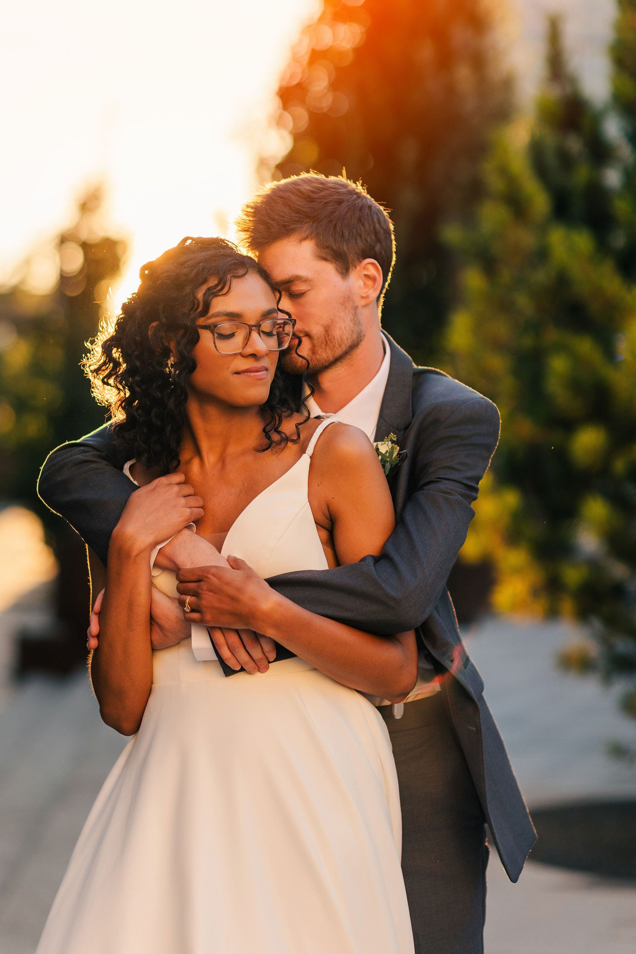 A couple in wedding attire embracing outdoors during sunset, with trees in the background.