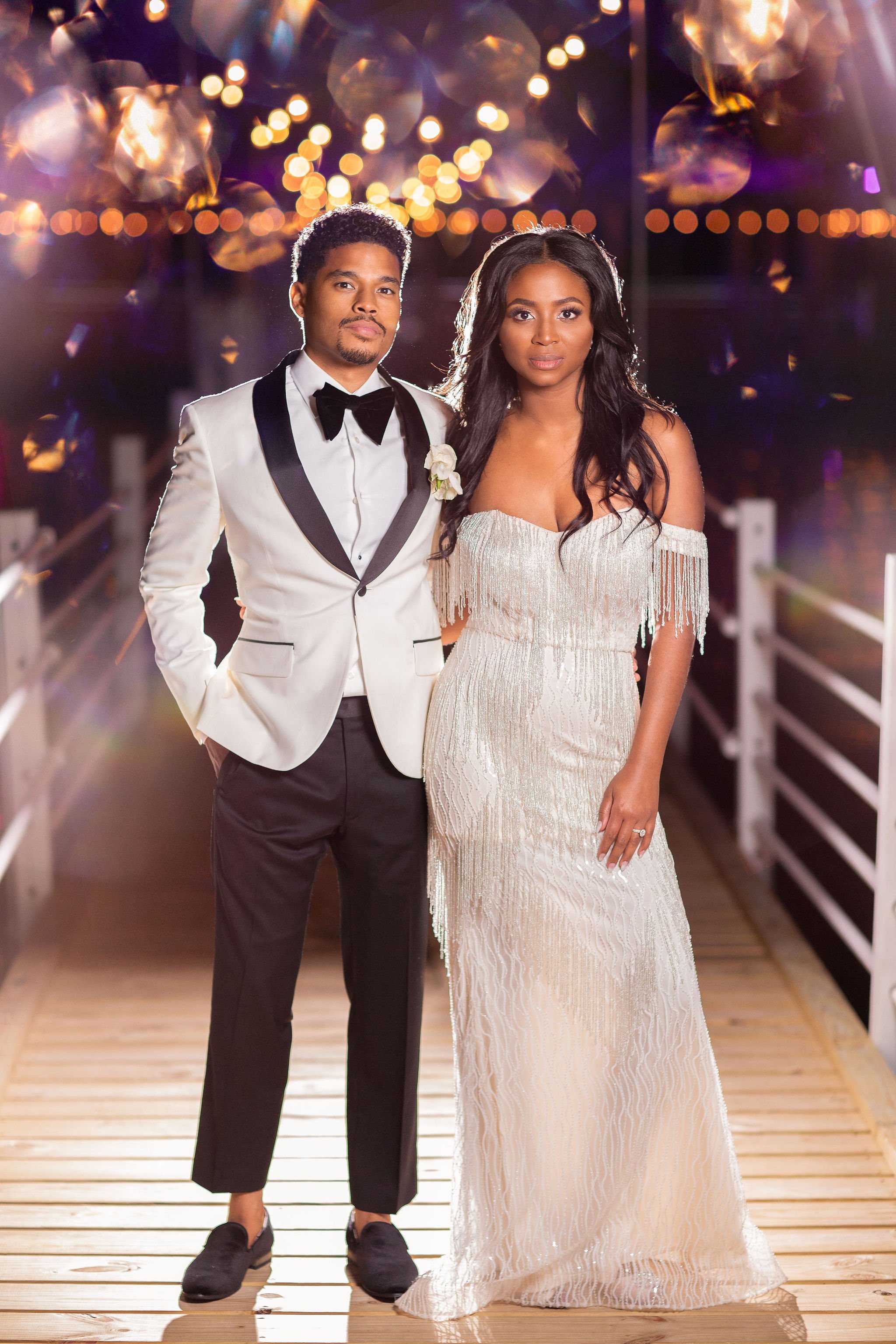A couple dressed in wedding attire standing on a wooden bridge at night with blurred lights in the background.