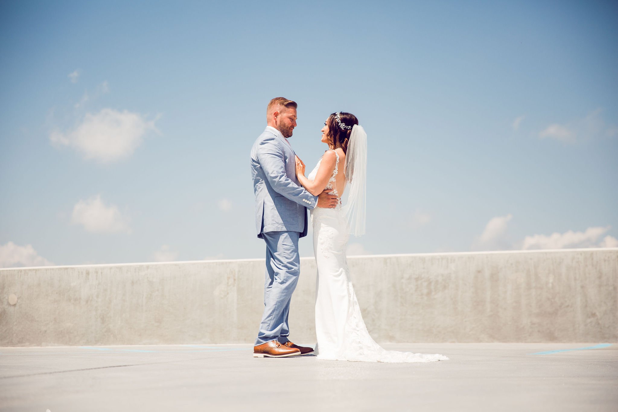 A bride and groom standing on a rooftop parking lot during their wedding, facing each other and holding hands, with a blue sky and a few clouds in the background.