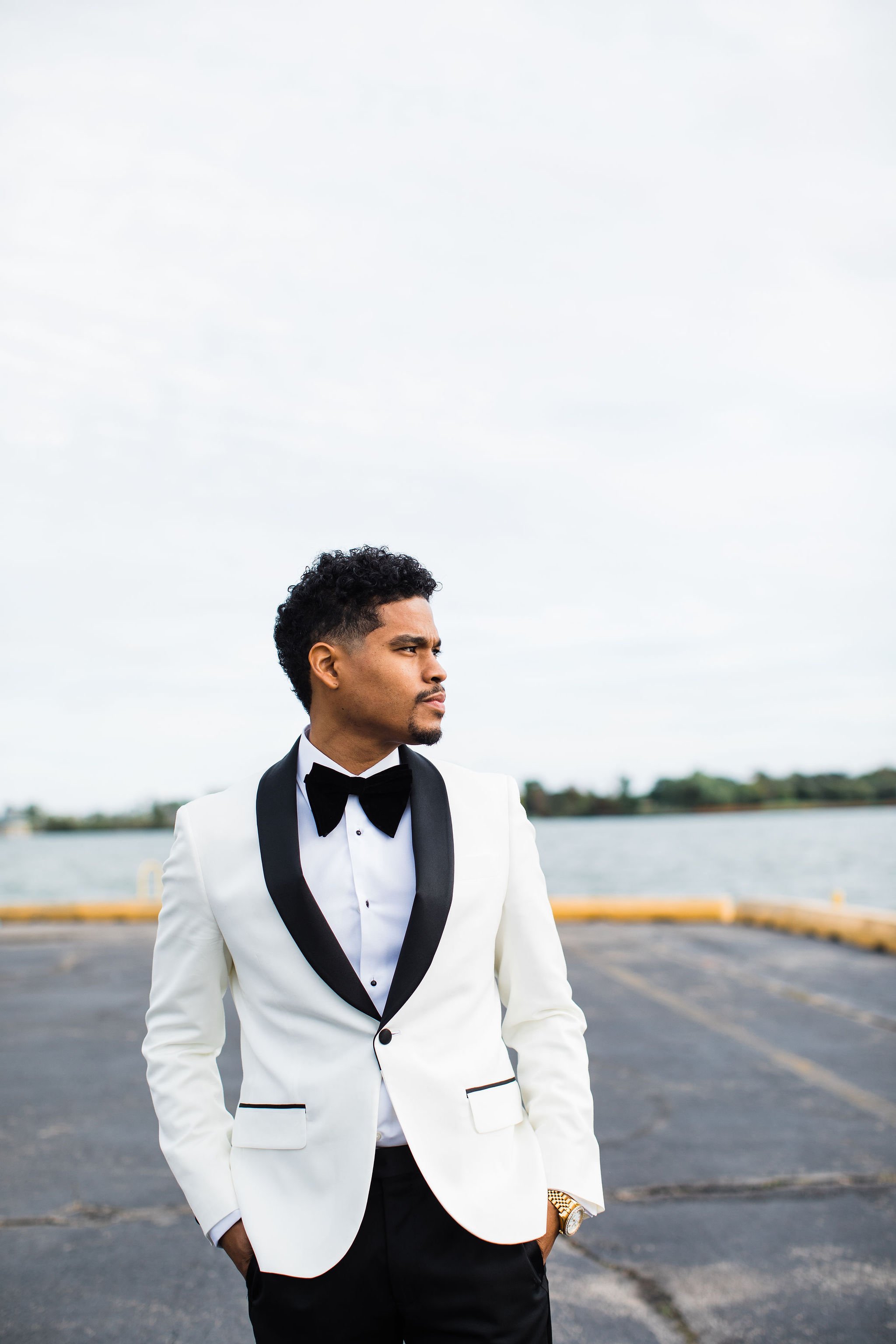 A man in a tuxedo with a white jacket, black bow tie, and black pants standing outdoors near water, looking to the side.