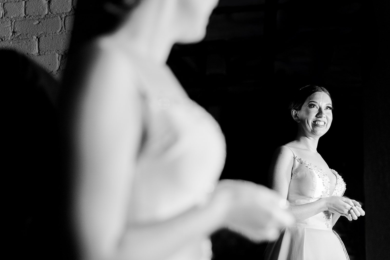 Black and white photo of a woman smiling and looking to the side, wearing a dress with lace details. Her hair is neatly styled, and she is standing against a dark background.
