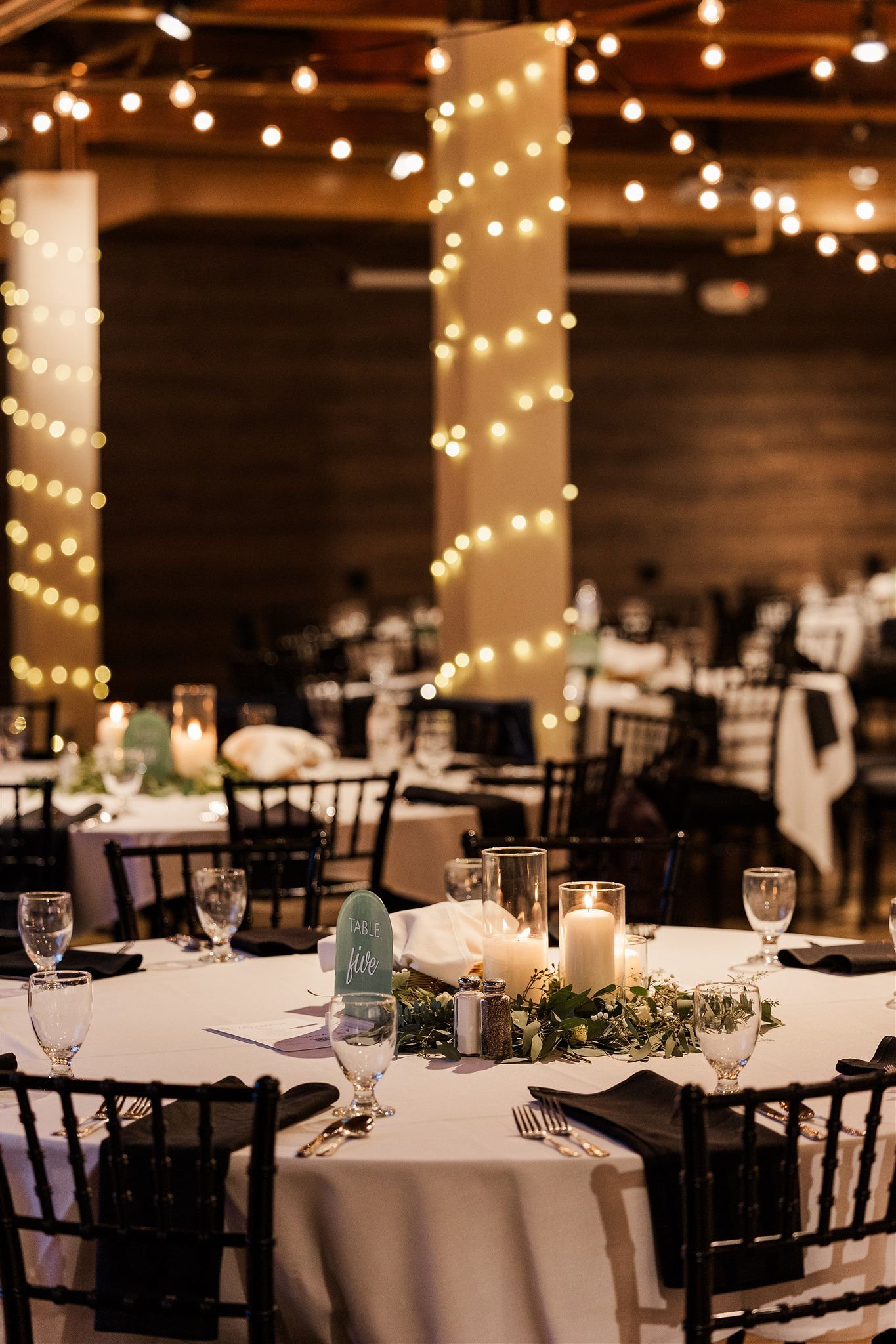 Elegant banquet table decorated with candles, greenery, wine glasses, and black napkins, surrounded by black chairs, with string lights hanging from the ceiling in a warmly lit venue.