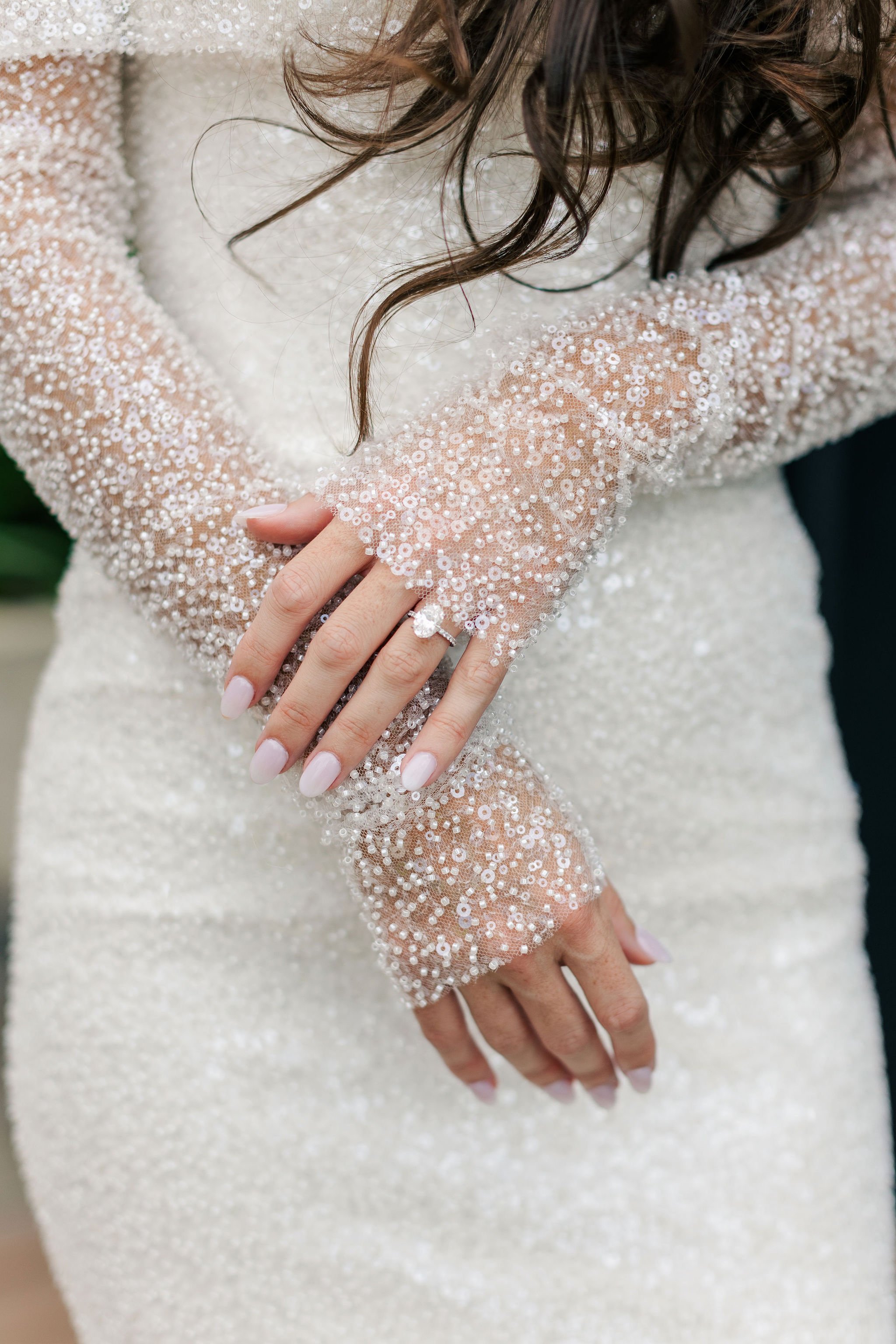 Close-up of a bride's hands with a wedding ring, wearing a long-sleeve dress embellished with white beads and sequins.