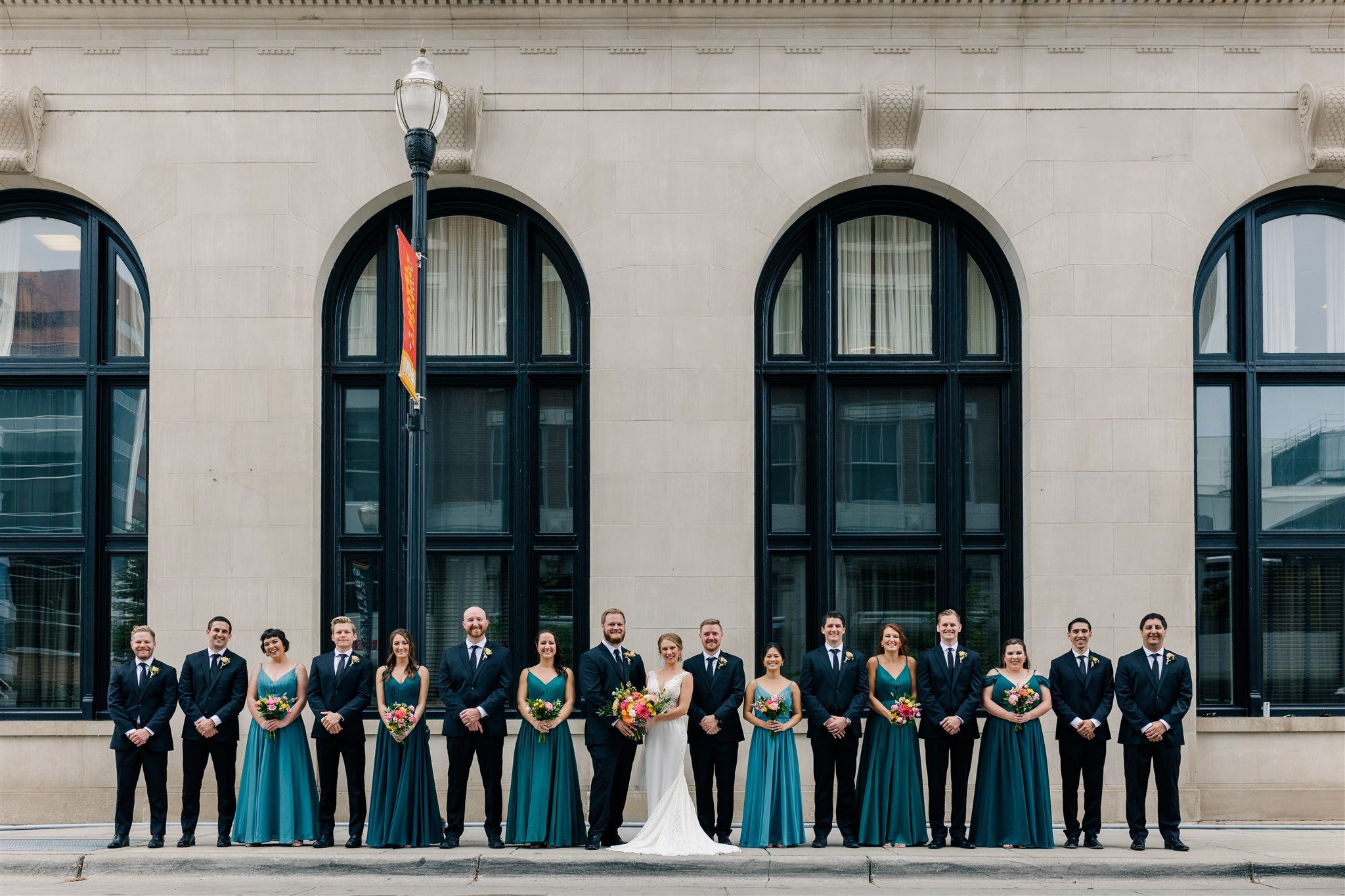 A wedding party with six bridesmaids and six groomsmen standing in front of a building with large arched windows, holding bouquets and dressed in formal attire.