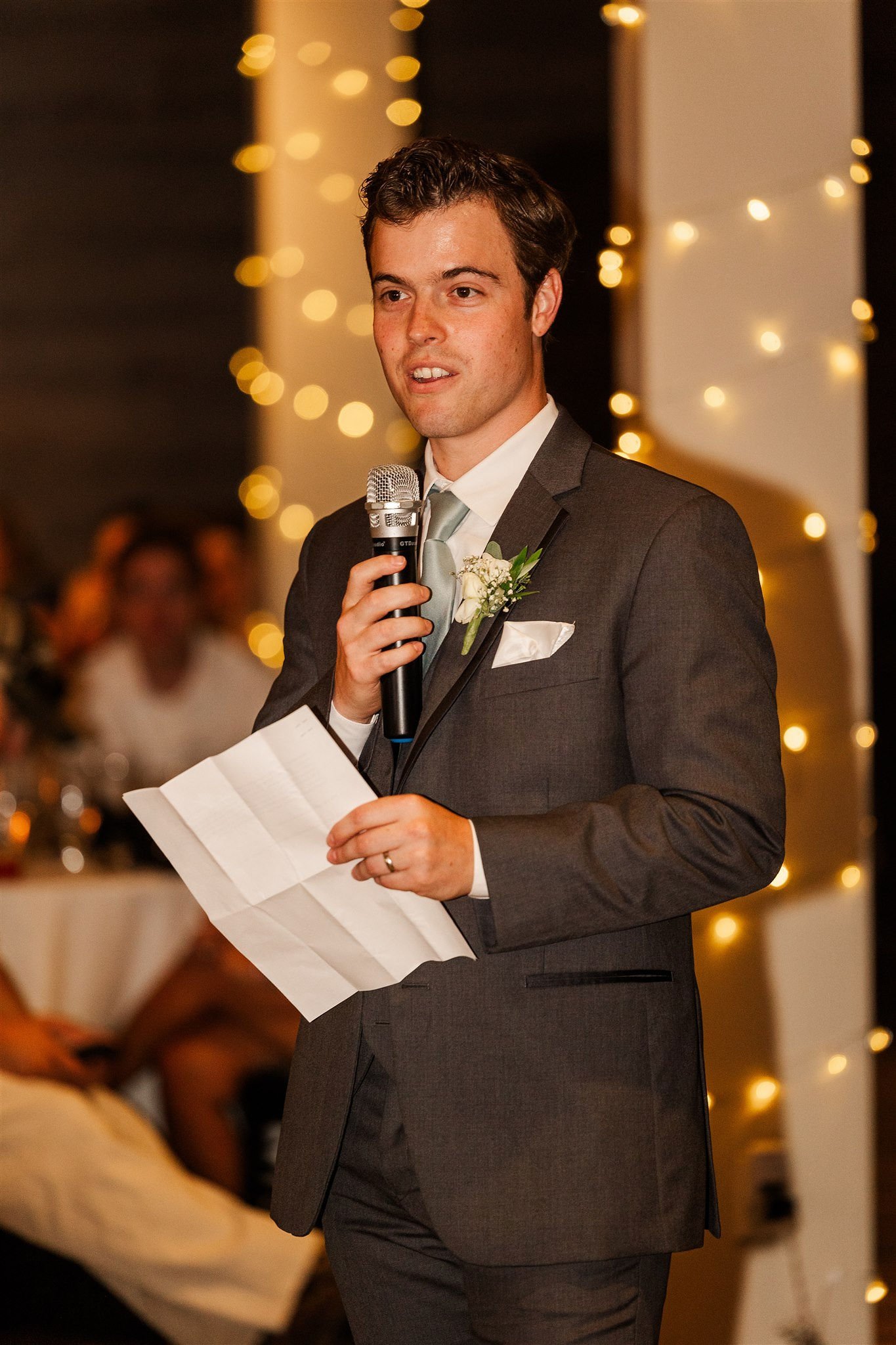 A man in a gray suit giving a speech at a wedding reception, holding a microphone in one hand and notes in the other, with a blurred background of guests and decorative string lights.