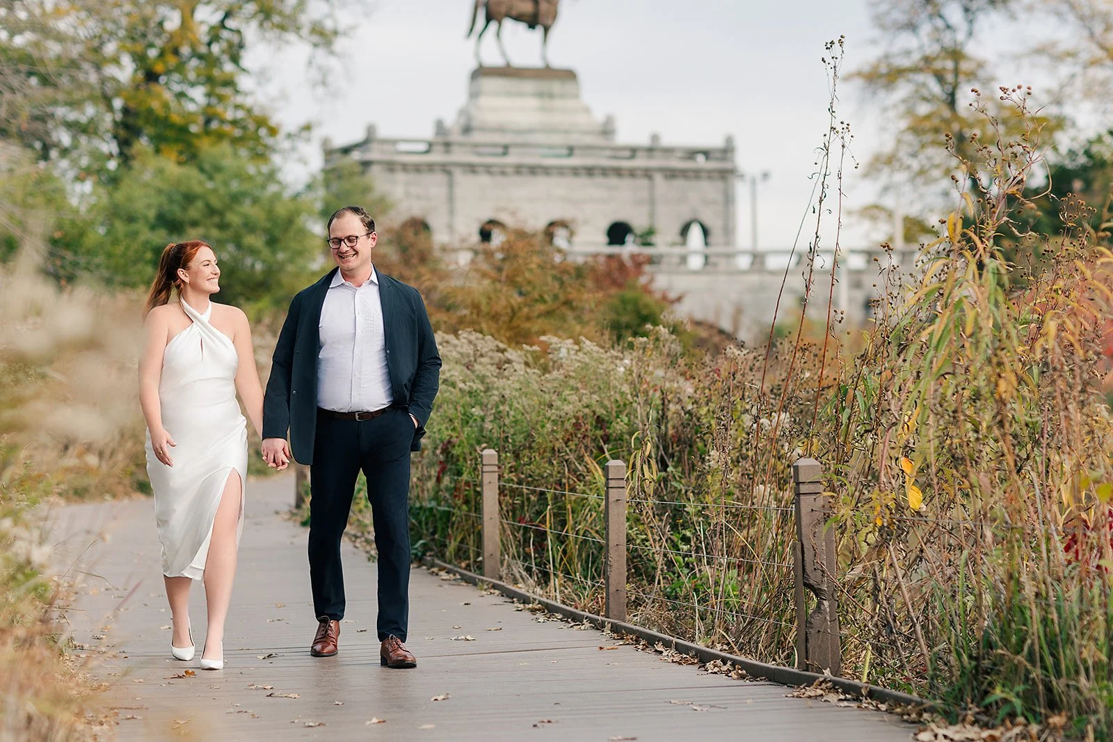 A couple holding hands and walking on a wooden path surrounded by fall foliage, with a historic monument in the background.