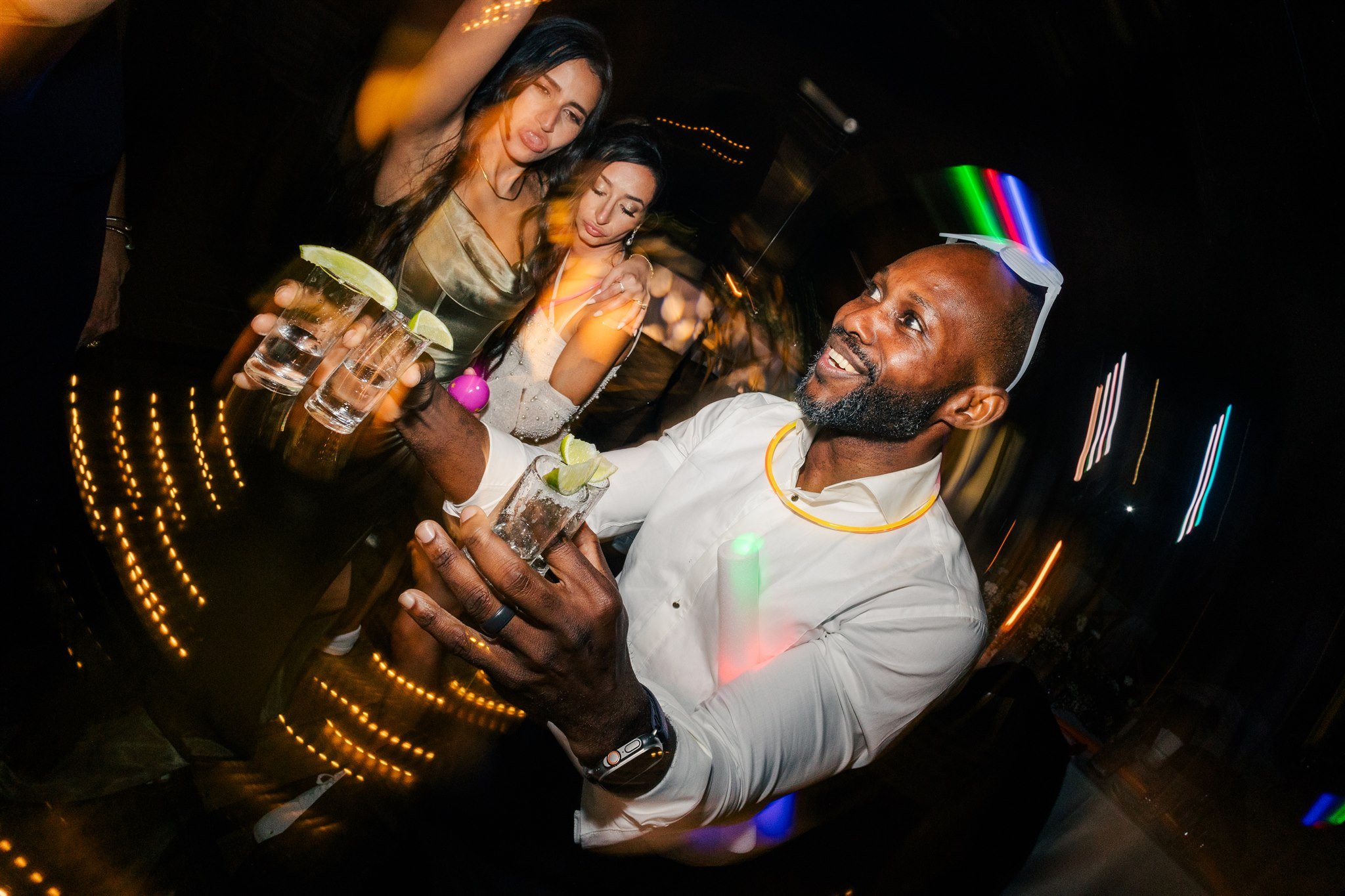 A bartender smiling and celebrating with women at a party, holding glasses with lime slices and drinks, with colorful lights in the background.